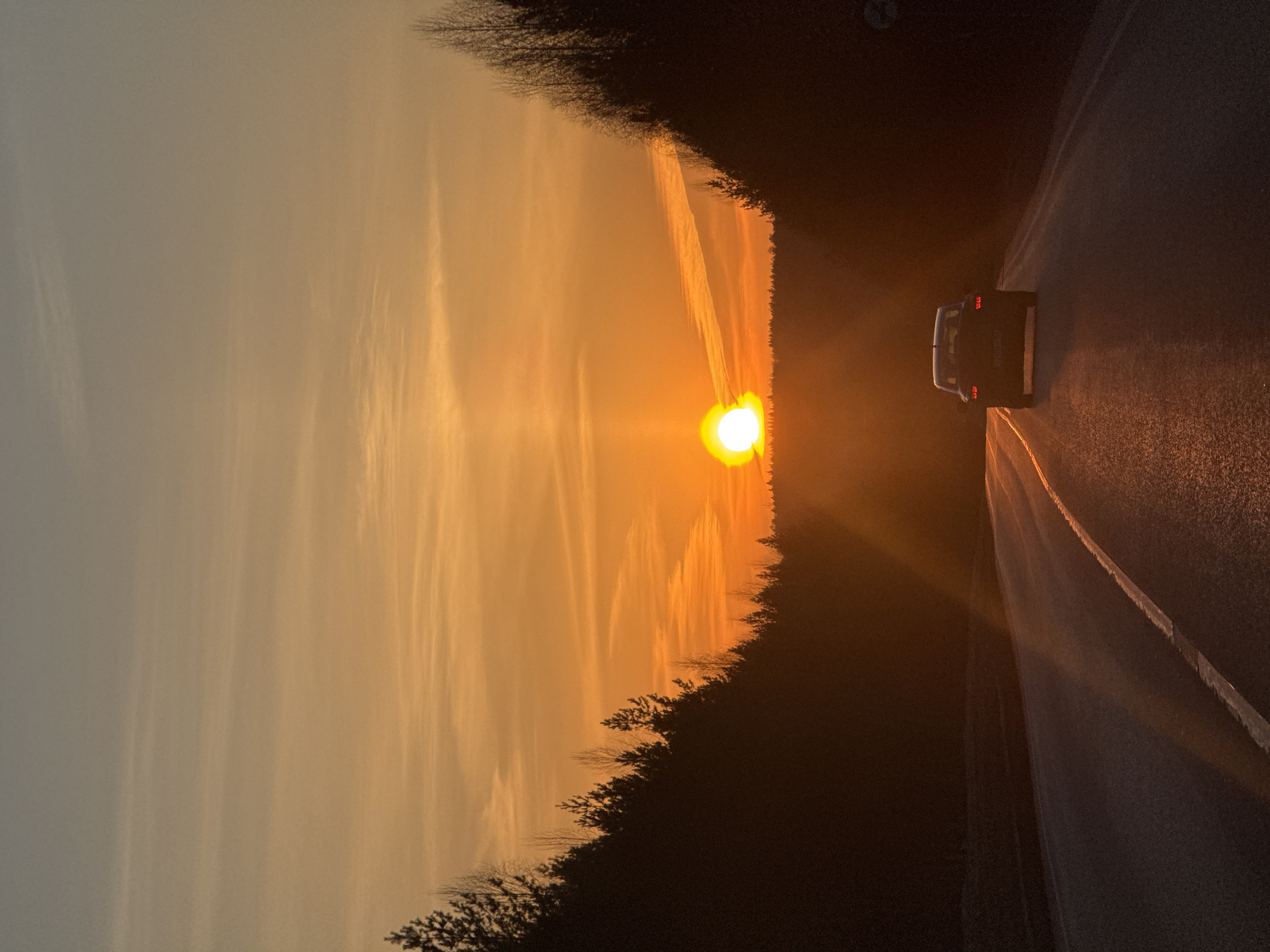 The image depicts a sunset over a road in Chaikovsky, Russia. A car is driving along the road, illuminated by its headlights and taillights. The sky is a gradient of orange and yellow, with streaks of clouds visible. The road is flanked by trees on both sides. The setting sun is positioned in the center of the image.