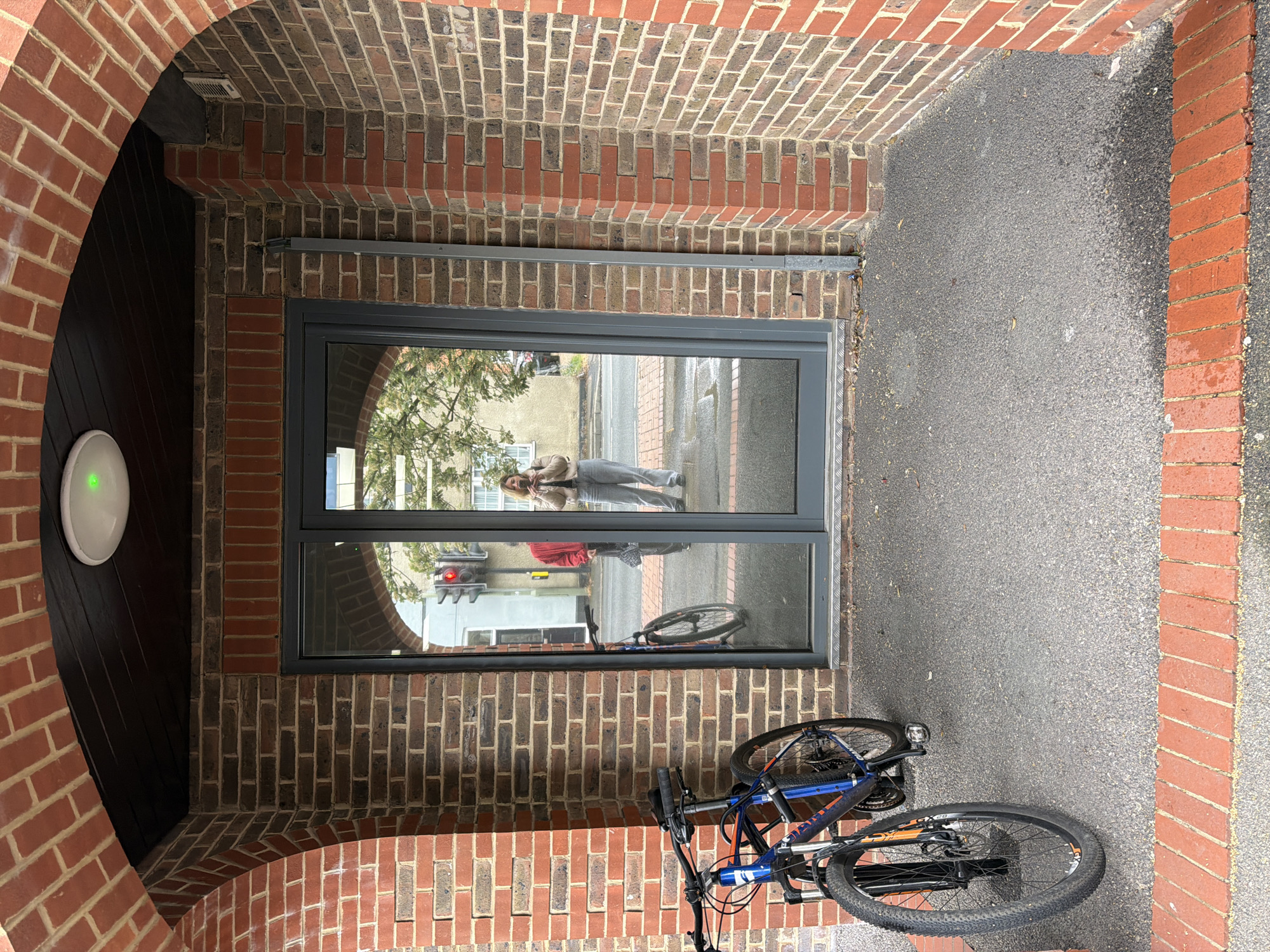 The image depicts the entrance to a building in Mole Valley, United Kingdom. The entrance is framed by a brick archway leading to a gray-framed glass door, which reflects a person taking a picture and a street scene with a traffic light. A blue and black bicycle is parked haphazardly in front of the door. The ground is paved with dark gray asphalt. A round, white light fixture is attached to the wall to the left of the door, showing a green light.