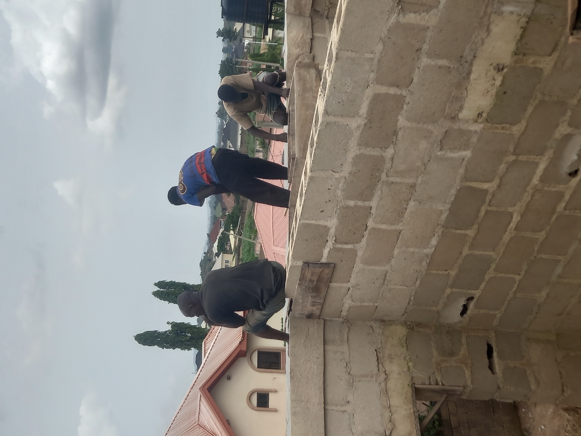In the city of Gura Top, Nigeria, three construction workers are seen on the rooftop of a partially built structure. One worker, wearing a dark t-shirt and shorts, is seated on the edge of the concrete wall, looking down and to his left. Another worker, dressed in a blue and red t-shirt and dark pants, is standing and bending over, facing away from the camera. The third worker, in a light brown t-shirt and shorts, is kneeling on the concrete and appears to be working with his hands. The construction site is characterized by cinder block walls and unfinished roofing. In the background, several houses with red roofs, tall trees, and a cloudy sky are visible, indicating a residential area within the city.