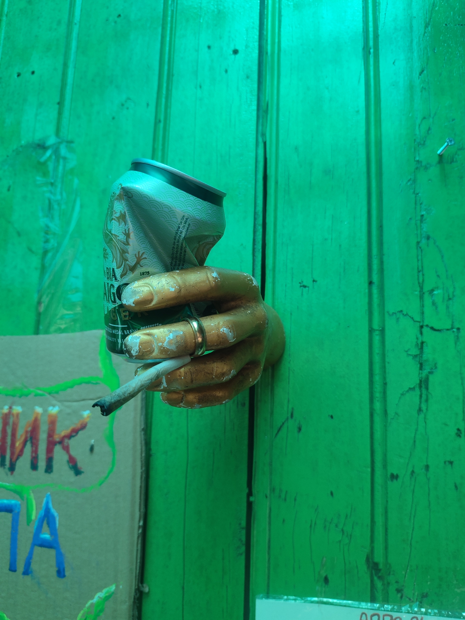On a weathered, vivid green wooden surface in what appears to be Cam Ly, Da Lat, Vietnam, a partially crushed can of "Bia Saigon" is held by a gold-painted plaster hand. The hand, adorned with a ring, holds a lit cigarette. Fragments of cardboard with Cyrillic script and blue writing are visible below the hand.