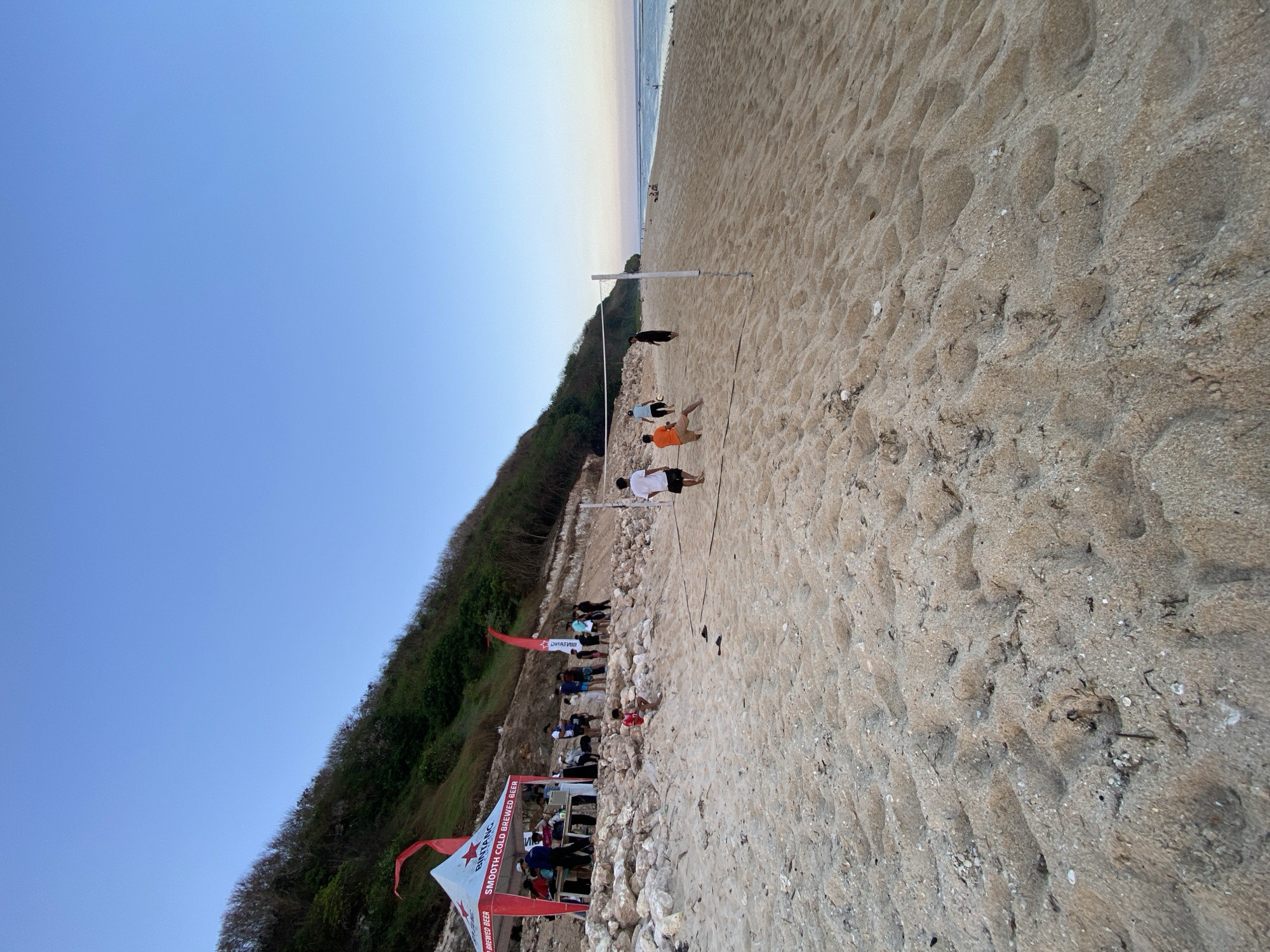 A wide-angle, low-angle shot captures a beach scene in Kuta Selatan, Indonesia. The foreground is dominated by the textured sand of a beach, with visible footprints and debris. In the mid-ground, a volleyball net is set up, and several individuals are engaged in a game or watching. A red and white tent with "BINTANG" branding and the slogan "SMOOTH COLD BREWED BEER" is visible on the left, with people gathered around it. Beyond the volleyball court, the ocean stretches out to the horizon, with a clear blue sky overhead. The background features a lush green hillside, indicative of the coastal landscape of Kuta Selatan. The lighting suggests it is late afternoon or early evening.