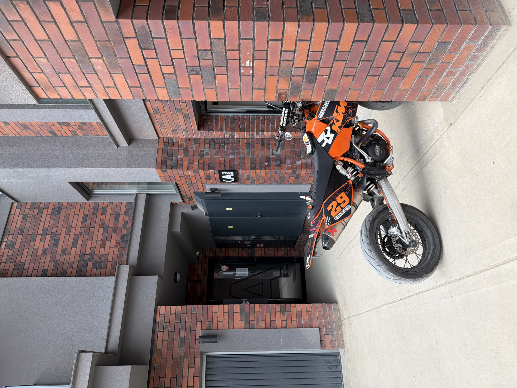An orange and black KTM motorcycle is parked on a light grey concrete surface in front of a modern building with a combination of red brick and grey concrete construction. The building includes a grey garage door at the bottom, followed by a doorway, a window, and a brick facade above. The motorcycle has a number "29" on its side. The photo is taken at an unusual angle, tilting the scene and making it appear as if the building is above the motorcycle. No people are visible in the image. The location is presumably in an urban setting within Sofia, Bulgaria, given the building's architectural style and materials.