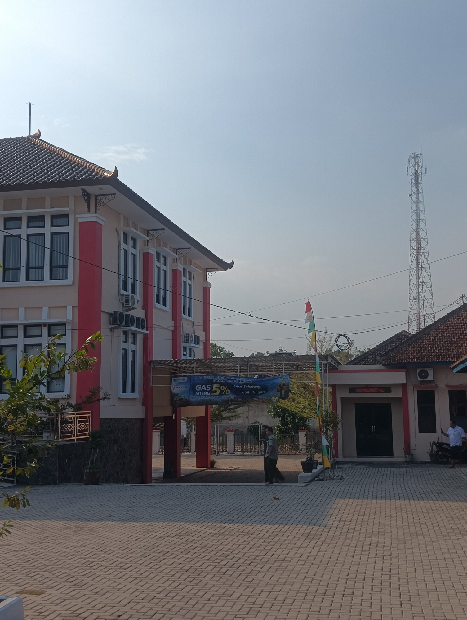 The image shows a building with a red and beige facade, a tiled roof, and several windows. In front of the building is a paved courtyard. A man in a white shirt and black pants is standing near the entrance, holding a smartphone. Another man in a gray shirt and black pants is walking away from the building. A red and white flag is visible on a flagpole. In the background, there is a tall telecommunications tower. The sky is clear and blue. The building appears to be a public or administrative building, possibly a government office or a bank, judging by the banner that reads "GAS JATENG 5%" which likely refers to a gas or utility service promotion in the Central Java region of Indonesia. The overall impression is of a sunny day in an urban or semi-urban environment in Indonesia.