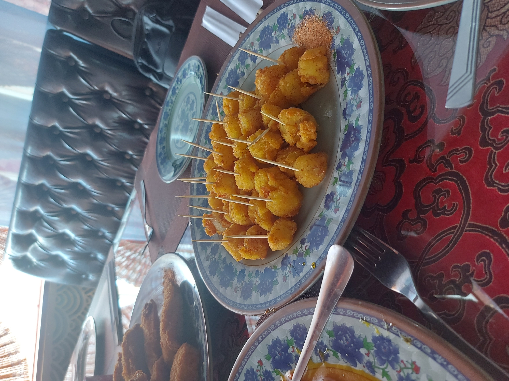 A close-up, eye-level shot shows two plates of fried food and dining utensils on a patterned red tablecloth. The plate in the foreground holds numerous bite-sized pieces of fried food, each skewered with a toothpick. A small pile of reddish-orange seasoning is visible to the right of the food. The plate has a blue and white floral pattern along its rim. To the left, another plate is partially visible, showing more fried food, possibly fish sticks. A fork is placed on the tablecloth to the left of the foreground plate. In the background, a dark, tufted leather couch is visible, along with a window with blinds, suggesting an indoor dining setting. The image was taken in Blagoveshchensk, Russia.