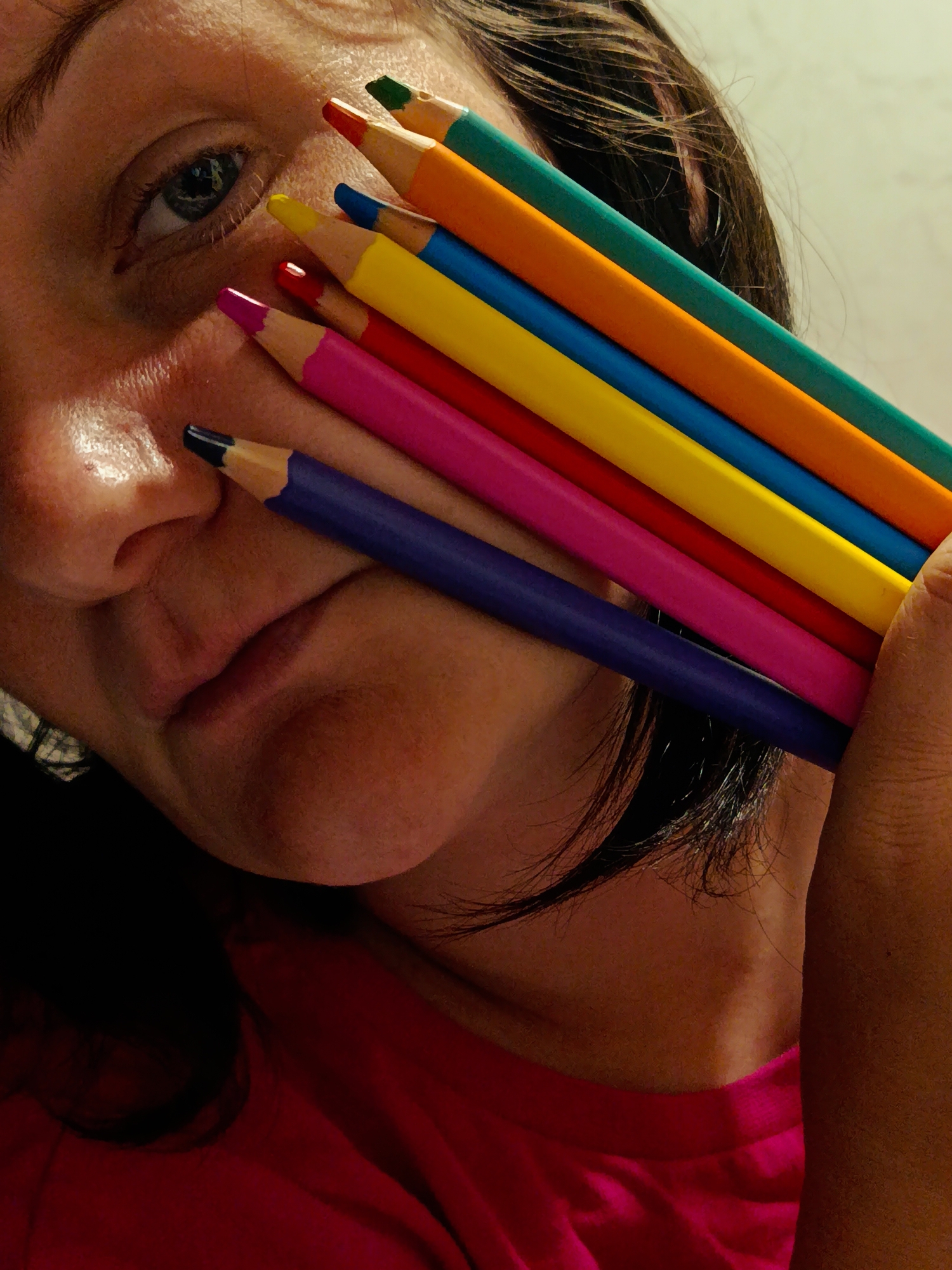 A close-up indoor shot shows a person holding a stack of colored pencils against their face. The person's eye is visible to the left, and the nose and mouth are partially obscured by the pencils. The pencils, from bottom to top, are dark blue, pink, red, yellow, light blue, orange, and green. The person is wearing a pink shirt. The background appears to be a plain wall. The location, Фоки, Russia, is not apparent from the visual content of the image.