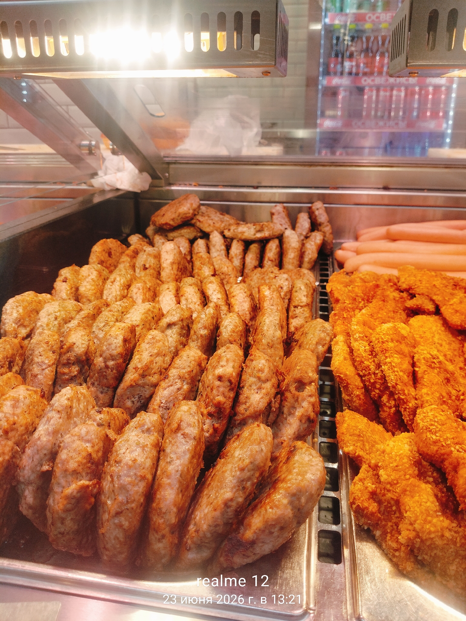 This media file shows a close-up of a refrigerated display case filled with various prepared food items. The primary subjects are food items, specifically what appear to be sausages, meatballs, and breaded chicken pieces. The food is arranged in metal trays within a well-lit commercial setting, likely a food service establishment such as a deli, supermarket, or fast-food restaurant.
In the foreground, a large quantity of cylindrical, brown, seasoned meat items, possibly sausages or meatballs, are neatly arranged. To the right, a section of the display holds golden-brown, breaded food items, likely chicken wings or tenders. In the background, behind a glass partition, a stack of pale pink sausages is visible. The setting is sterile and functional, emphasizing the food presentation. Above the display, fluorescent lights cast a bright, warm glow on the food and the metallic interior of the unit. In the far background, through another pane of glass, shelves stocked with bottled beverages, predominantly Coca-Cola, are visible.
The image captures a moment of food display, suggesting it's during operating hours, likely daytime given the bright lighting. There are no people visible in the frame. The overall impression is one of abundance and readily available prepared food.
Visible text includes "realme 12" in white and "23 ИЮНЯ 2026 г. В 13:21" in white, which appears to be a date and time stamp, indicating June 23, 2026, at 13:21. Other partial text is visible on the beverage bottles in the background, which appears to be Cyrillic lettering for "ОСВЕЖИТ" (Osvyezhit), meaning "refresh."
