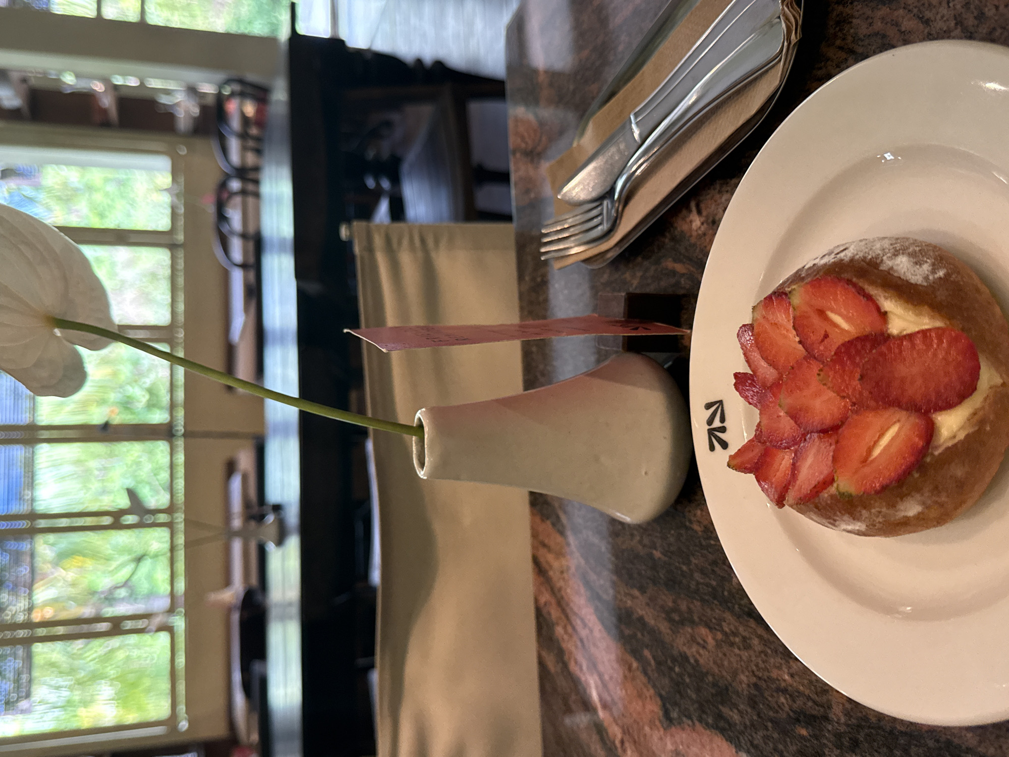 This is a vertically oriented photograph, providing a slightly angled, close-up view of a table setting in what appears to be a cafe or restaurant in North Kuta, Indonesia.
In the foreground, a white ceramic plate holds a light brown pastry or bun, generously topped with thinly sliced fresh strawberries and a dollop of white cream. The pastry is dusted with powdered sugar. Next to the pastry, on the white plate, a black stylized logo resembling the letters "RZ" or a similar symbol is visible.
To the left of the plate, a small, elegant white ceramic vase holds a single white anthurium flower with a long green stem. Tucked into the vase, or perhaps attached to it, is a small, rectangular red/pink card, possibly a label or decorative element, though no specific text is legible on it.
Further back on the table, a set of polished silver cutlery (a fork and a knife) rests on a folded brown napkin or paper wrapper. The table surface beneath these items is a dark, mottled material, likely granite or marble, featuring shades of brown, black, and reddish-brown.
The background shows a brightly lit interior space with large windows revealing lush green foliage outside, suggesting a tropical or garden setting and indicating daytime. The window panes create a grid pattern, and the light through them produces soft bokeh effects. Furniture and indistinct elements of the cafe interior are visible through the window's reflection and beyond. The overall ambiance is tranquil and inviting.