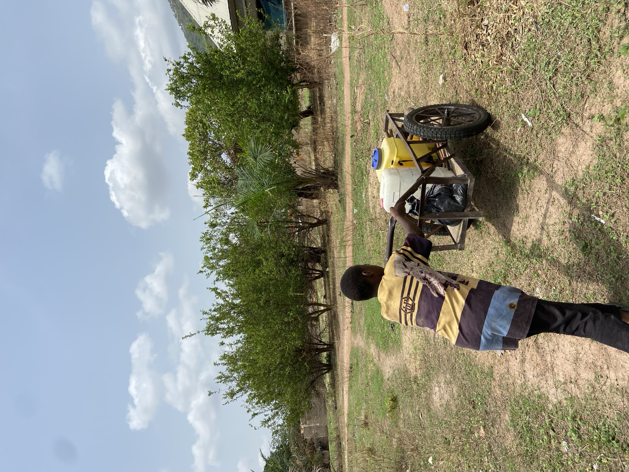 A young person stands on a dirt path in Mararaba, Nigeria, pulling a wheeled cart. The cart carries two plastic containers, one yellow and one white, and a black plastic bag. The person wears a striped shirt and black pants. Trees line the path to the left. The sky is partly cloudy. The ground is covered with dry grass and dirt.