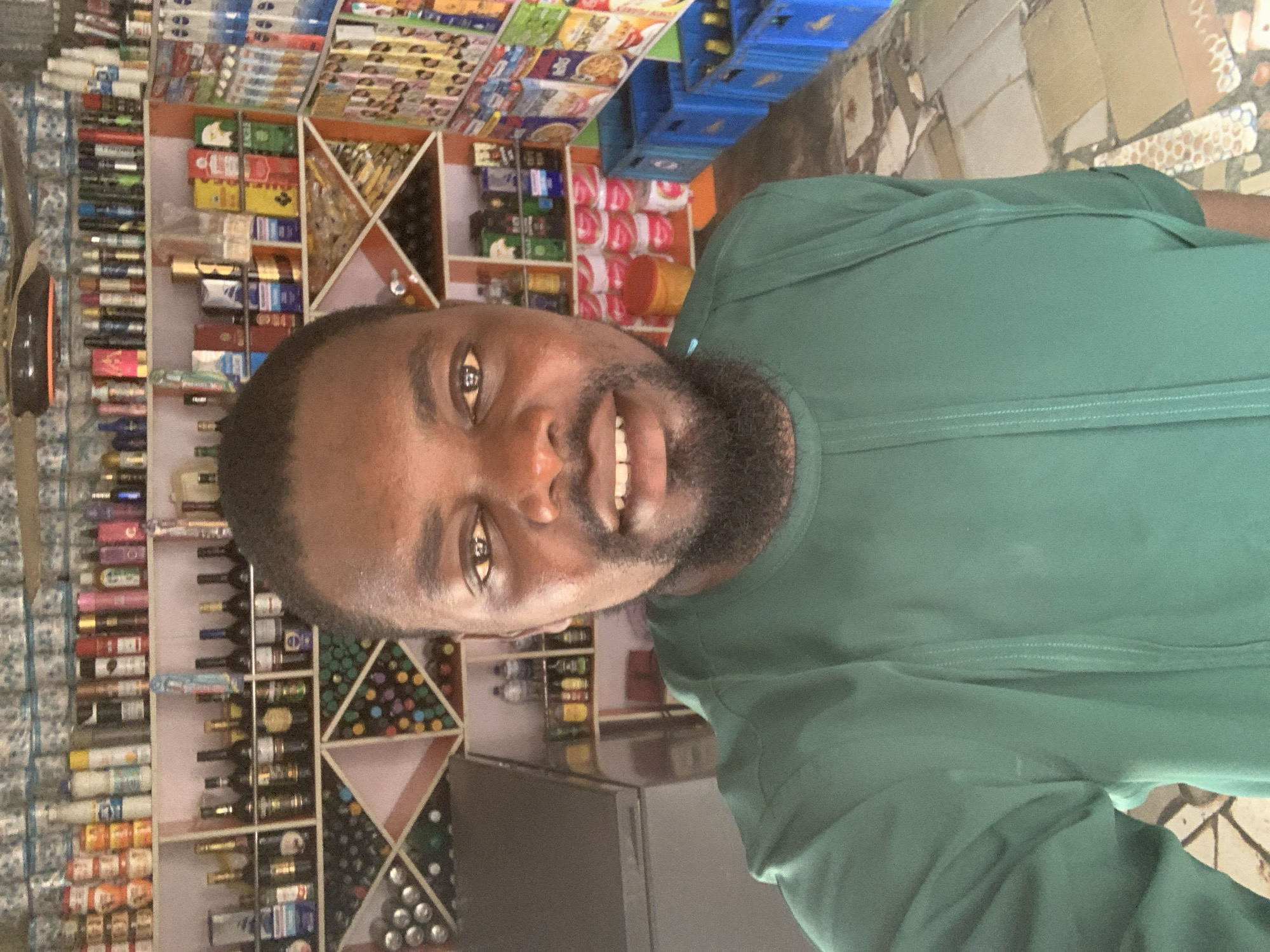 A medium shot selfie shows a Black man in a green tunic smiling at the camera. He is standing in a small shop filled with shelves stocked with various items, including bottles of alcohol, canned goods, and toiletries. The shop appears to be located in the city of Shagamu, Nigeria. The man's face is well-lit, with his eyes open and his mouth slightly ajar, revealing his teeth. He has a short beard and short, dark hair. Behind him, shelves are filled with a variety of products, including what appear to be alcoholic beverages, perfumes, and canned foods. The shop's interior is decorated with colorful patterns on the walls and shelves. The floor is tiled with various shades of brown and gray. The overall impression is that of a well-stocked convenience store.