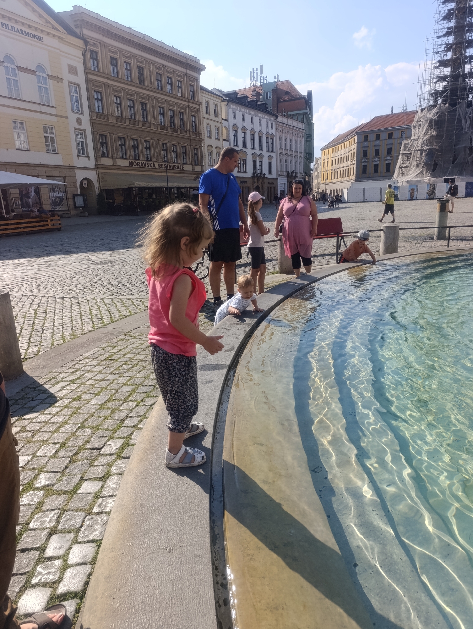 The image captures a scene in the city of Olomouc, Czechia, likely in the vicinity of a fountain. In the foreground, a young girl with blonde hair is standing by a fountain, wearing a pink sleeveless shirt and patterned leggings. Beside her, a baby is crawling at the edge of the fountain. Further back, a woman in a pink dress stands near a young girl in a pink hat and white shirt. A man in a blue shirt and dark shorts is also visible. The background reveals urban architecture, including buildings with traditional facades and a scaffolding-covered structure, possibly a tower undergoing renovation. The ground is paved with cobblestones, and the water in the fountain is clear and reflects sunlight.
