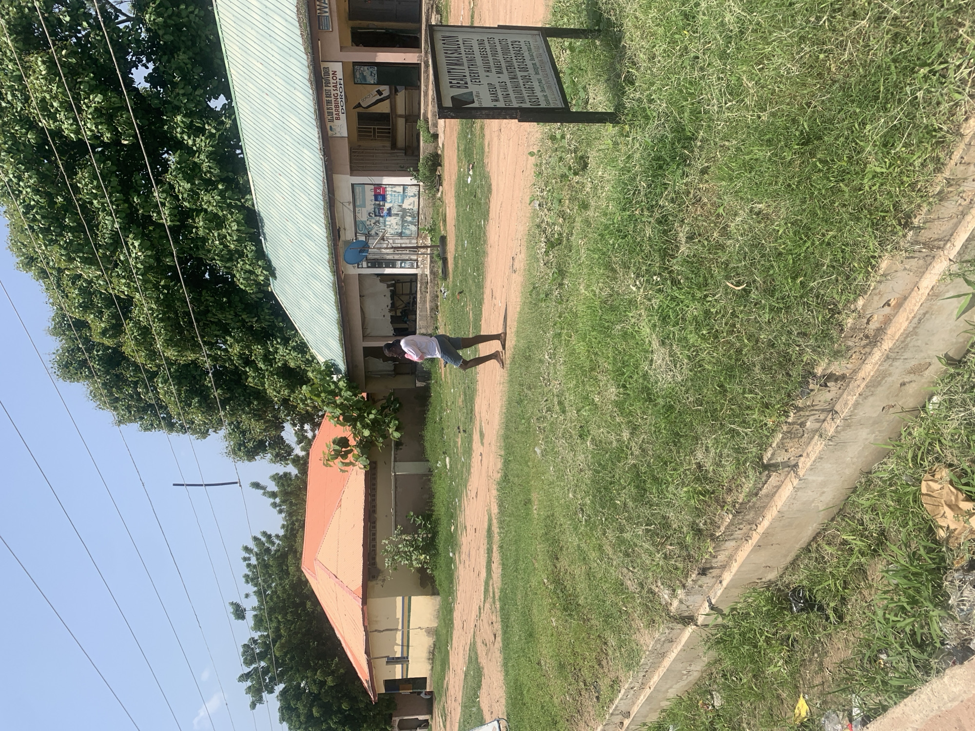 The image depicts a street scene in Jalingo, Nigeria. In the foreground, a concrete drain runs along a grassy area littered with trash, including plastic bags and paper. The ground is a mix of dirt and grass. Behind the drain, a dirt road leads past several buildings. To the left, a single-story building with a yellow facade and an orange, corrugated metal roof is visible. Further down the road, to the right, is a longer, single-story building with a green, corrugated metal roof. This building appears to house businesses, with signage visible above the storefronts. One sign reads "ALLAH IS THE BEST PROVIDER BARBING SALON DOROFI". Another sign, prominently placed in the middle ground, is for "BEAUTY MAX SALOON". It lists services such as makeup, hairdressing, fixing nails, and makeup products, along with phone numbers. A young person, wearing a white t-shirt and denim shorts, walks across the dirt road in the middle ground, facing away from the camera. The sky is clear and blue, and a large tree with dense green foliage is situated behind the buildings. Power lines are visible strung across the sky.
