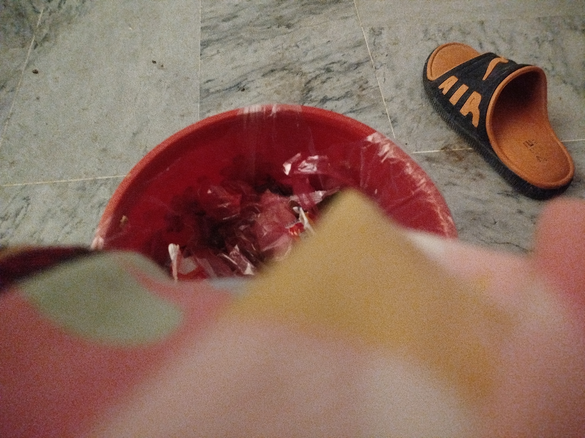 A high-angle, close-up shot captures a red plastic bucket on a marble floor. Inside the bucket, there appears to be crumpled plastic wrap, possibly containing some items, though they are not clearly discernible. To the right of the bucket, a black and orange slide sandal is positioned on the floor. The sandal has the word "AIR" printed in orange across the strap. The foreground is dominated by a blurry, out-of-focus area of pink, yellow, and green hues, suggesting a hand or an object is very close to the camera. The background shows a continuation of the marble flooring. The scene appears to be indoors, with no specific urban or rural context evident from the provided image.