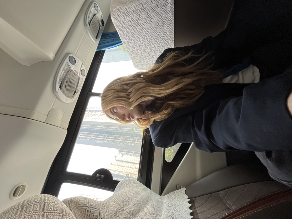 The image displays the interior of a vehicle, likely a bus or coach, positioned within an urban environment consistent with the Yanta District, China, as indicated by the modern high-rise buildings visible through the windows. A young woman with long, wavy blonde hair is seated on the right side of the frame, captured in profile. She is looking out the window to her left, wearing a dark blue oversized blazer over a light-colored inner garment. The vehicle's interior features white ceiling panels containing integrated overhead lights and speaker grilles. The seats are upholstered in a dark material, with light-colored, intricately patterned fabric covers on the headrests. A blue curtain is partially visible on the right side of the frame.