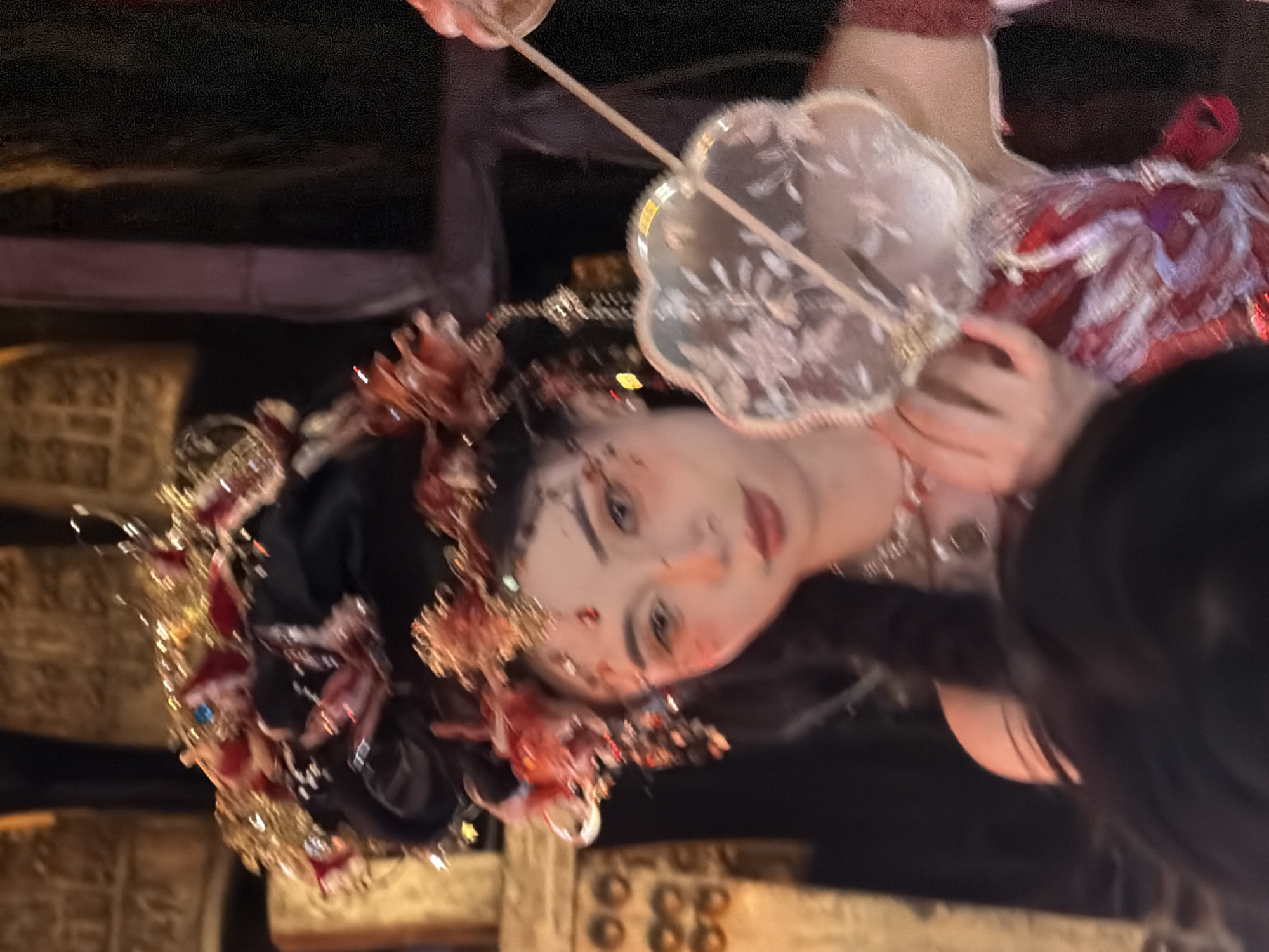 In Yuzhong District, China, a woman with ornate headdress and makeup, possibly a performer, holds a decorative fan. The image captures her face and upper body, with a blurred foreground suggesting an audience or other participants. The background is dimly lit and indistinct, with hints of gilded structures or artifacts. Her attire includes a red and white patterned garment and a jeweled headpiece adorned with red floral elements. The overall scene appears to be staged, perhaps for a cultural event or performance.