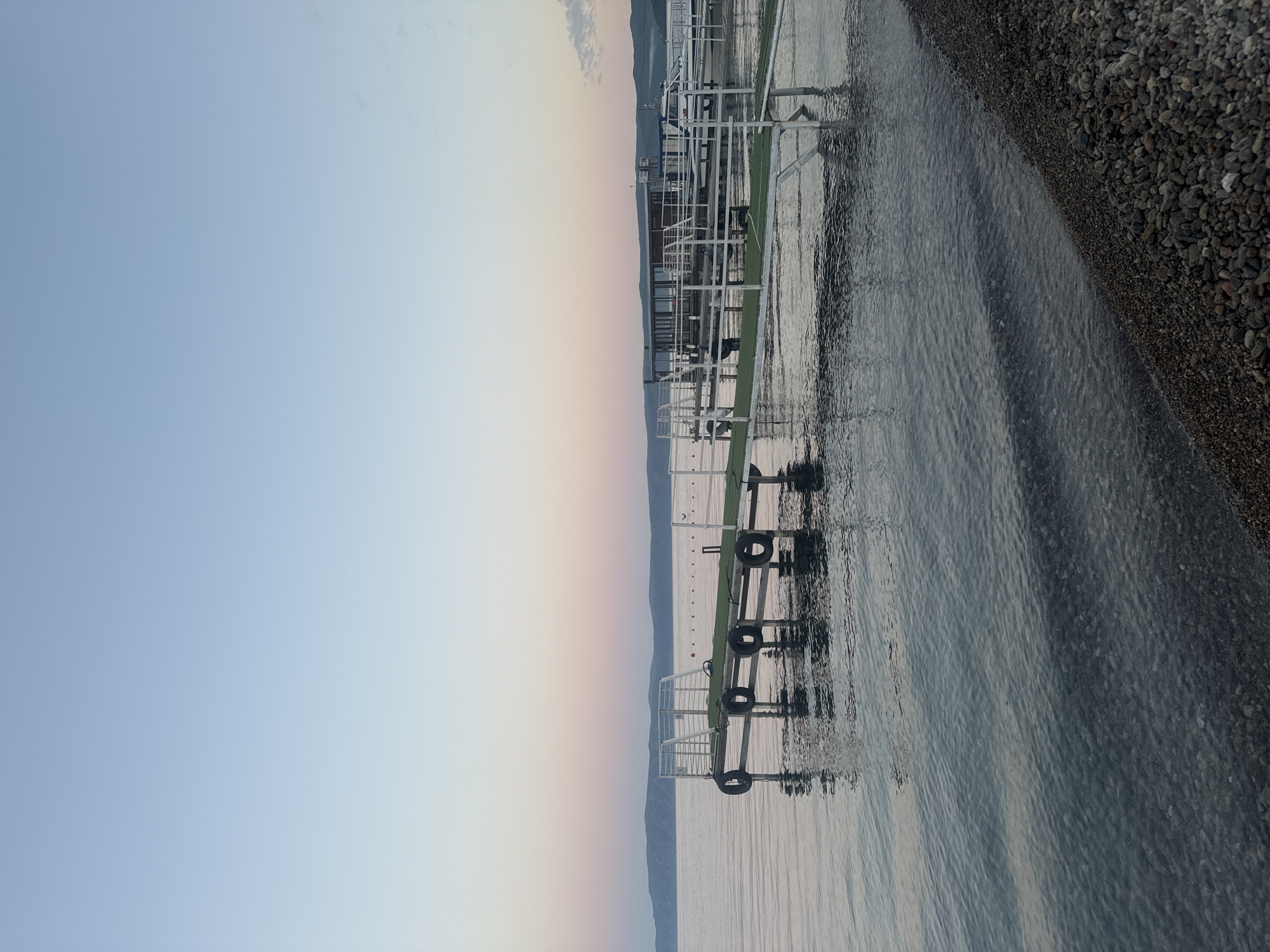 This is a landscape photograph taken at dusk on the shore of Lake Baikal in Listvyanka, Russia. The foreground shows a pebble beach with the clear, shallow water of the lake lapping at the shore. In the middle ground, a wooden pier extends into the lake. The pier is equipped with white railings and black tires used as fenders. Beyond the first pier, a larger, more complex pier structure is visible, featuring a building with glass windows and what appears to be a boat moored alongside. The water is calm and reflects the soft colors of the sky. In the background, rolling hills with a faint texture of trees can be seen along the horizon. The sky transitions from a pale blue at the top to soft pink and peach hues near the horizon, indicating the early stages of sunset or the end of twilight. There are no people visible in the image.