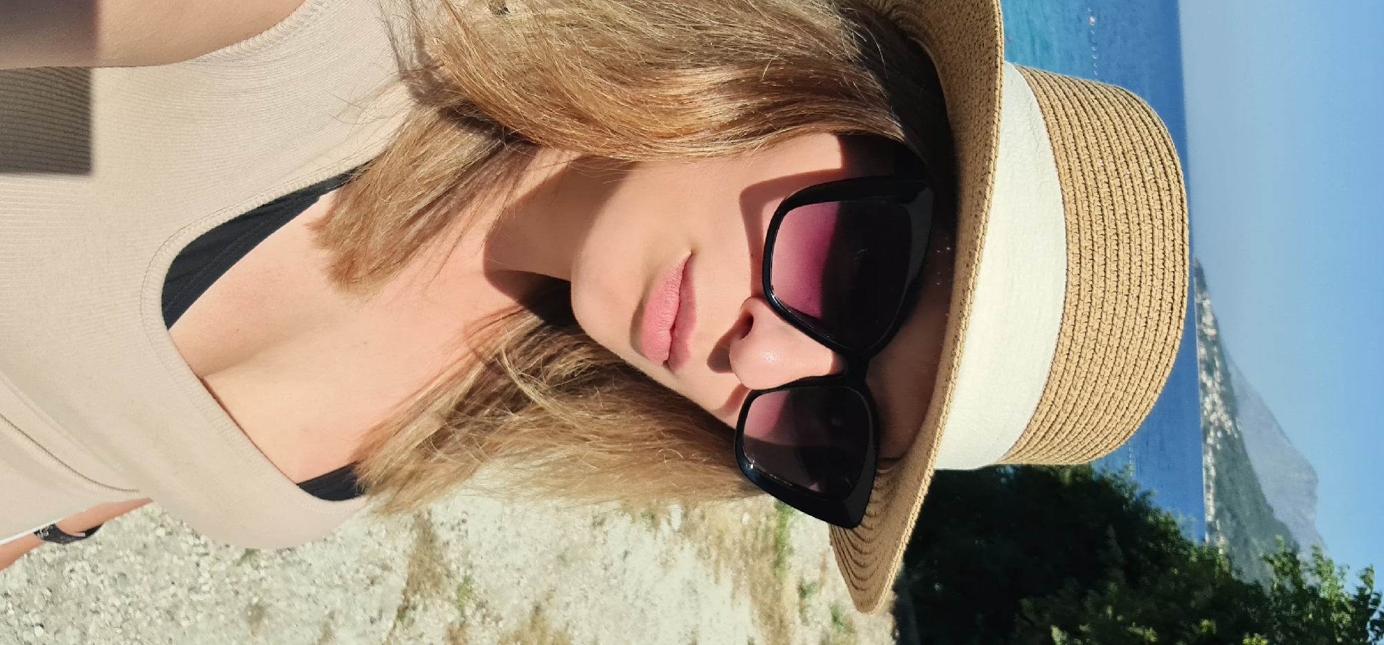 In the city of Općina Gradac, Croatia, a woman stands outdoors, facing the camera in a close-up shot. She is wearing a straw hat with a white band and large, black, cat-eye sunglasses. Her blonde hair is visible around her face. She is also wearing a beige, ribbed tank top, revealing a black bra underneath. The background shows the sea, a distant coastline with buildings, and lush green trees. The ground in the foreground is a light-colored gravel or sand. The lighting suggests it is a sunny day.