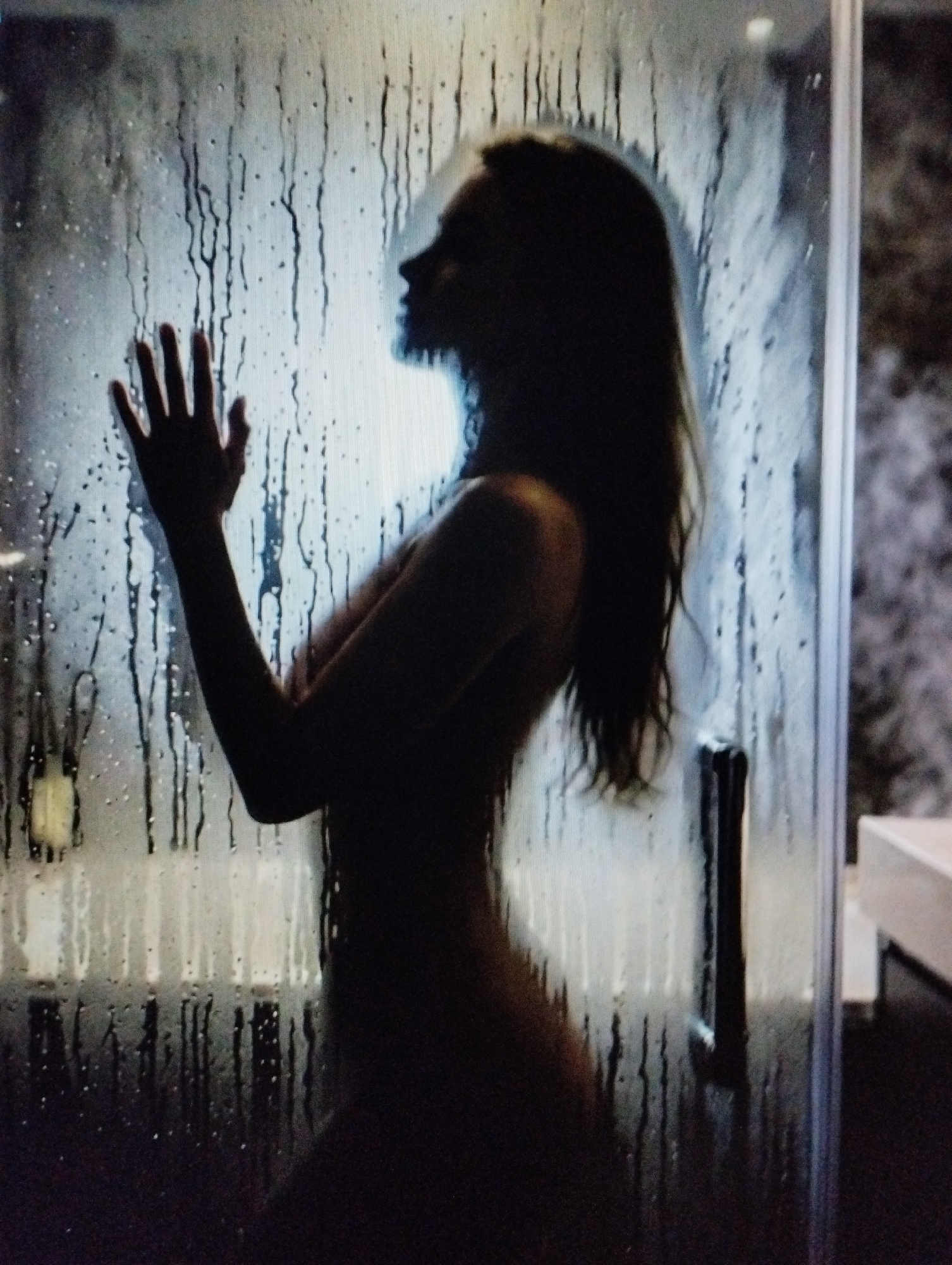 The image displays the silhouette of a person, likely female, standing behind a glass shower door that is covered in water droplets and streaks. The person's body is turned sideways, with their left hand pressed against the glass. Their head is tilted slightly, and their long hair cascades down their back. The lighting is dim, with the background appearing blurred and indistinct, suggesting an indoor setting, possibly a bathroom. The overall impression is one of privacy and intimacy.