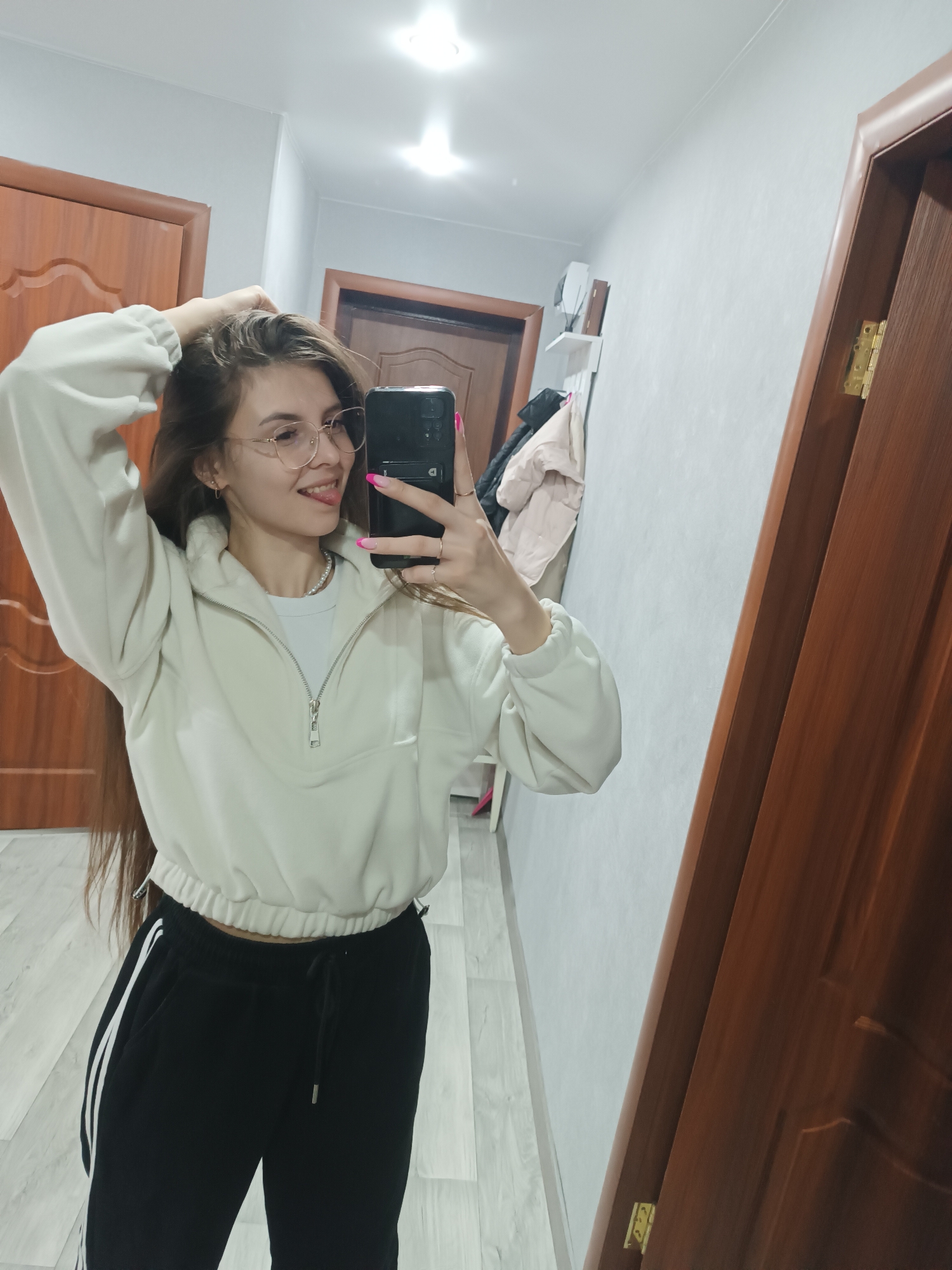 An indoor mirror selfie depicts a young adult female in a hallway or room within the city of Unknown, Unknown. The female, centered in the frame, has long brown hair and is wearing eyeglasses, a cream-colored quarter-zip sweatshirt over a white t-shirt with a pearl necklace, and black sweatpants with white side stripes. She smiles and extends her tongue while holding her long hair with her raised left hand. Her right hand holds a dark-colored smartphone, visible in the mirror's reflection, with her fingernails painted pink with light tips. The environment features light grey walls, light-toned wood or laminate flooring, and two paneled wooden doors, one on the left and one partially framing the mirror on the right, which shows brass-colored hinges. In the reflected background, a white shelf with various objects and hanging clothing is visible on the wall opposite the camera. Overhead, recessed circular lights illuminate the white ceiling.