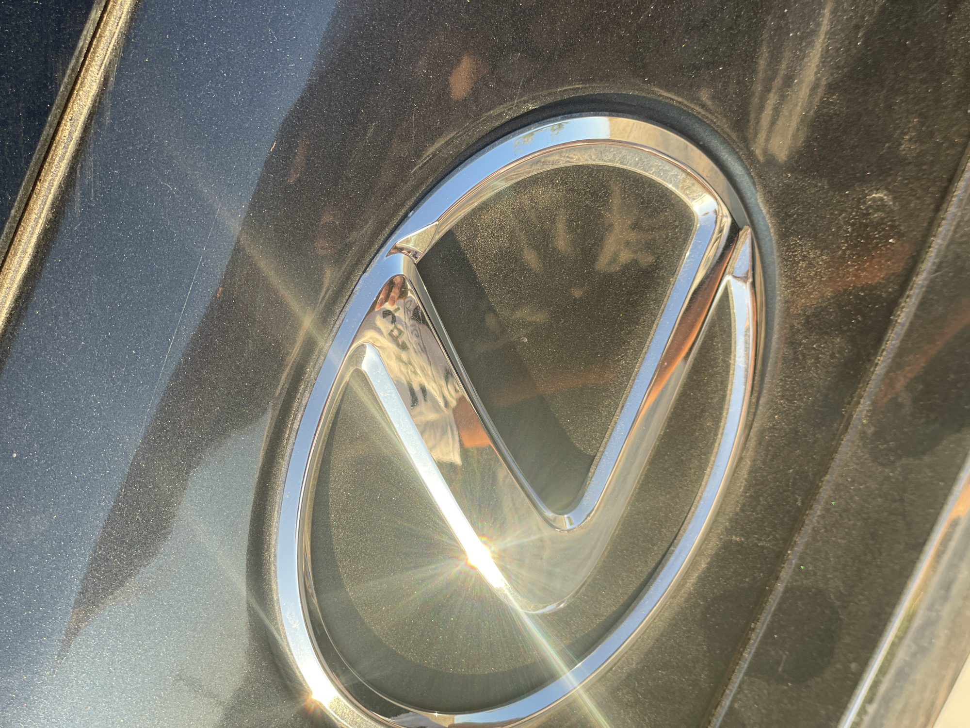 A close-up shot of the Lexus emblem on a dark-colored car is captured in the city of Aktau, Kazakhstan. The emblem, made of chrome, is oval-shaped with a stylized "L" inside. Sunlight reflects off the emblem, creating a starburst effect and illuminating the dusty surface of the car. The car appears to be parked outdoors, as evidenced by the natural lighting and the presence of dust and dirt on its exterior. The background is out of focus, but hints of a reflective surface, possibly pavement or a building, can be seen. The image is framed such that the emblem is the primary subject, occupying a significant portion of the frame.