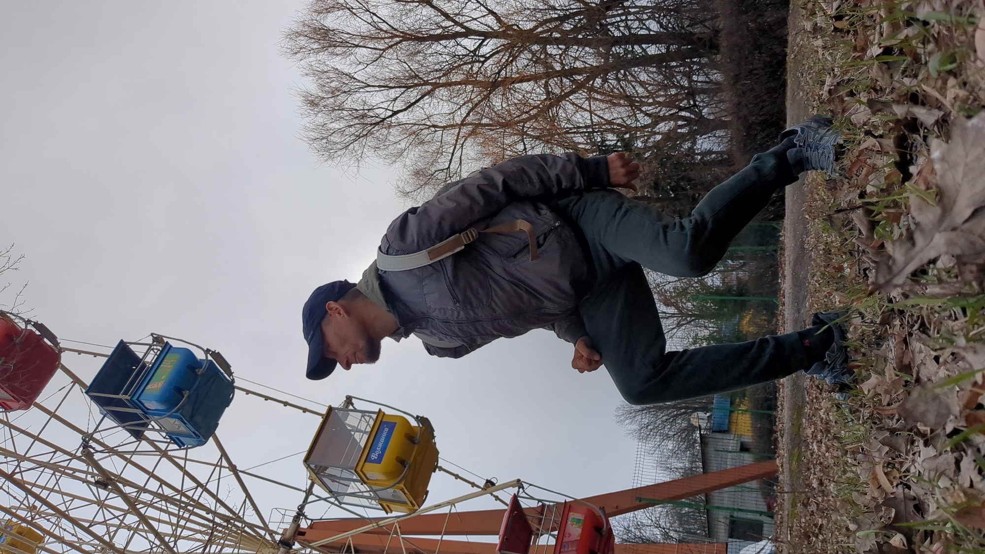 A man in dark clothing and a baseball cap appears to be falling upside down, with his legs oriented towards a ground covered in dry leaves, and his upper body leaning towards a Ferris wheel with multicolored cars. The Ferris wheel is partially visible against a gray sky, with bare trees in the background. The location is identified as Ulyanovsk, Russia.
