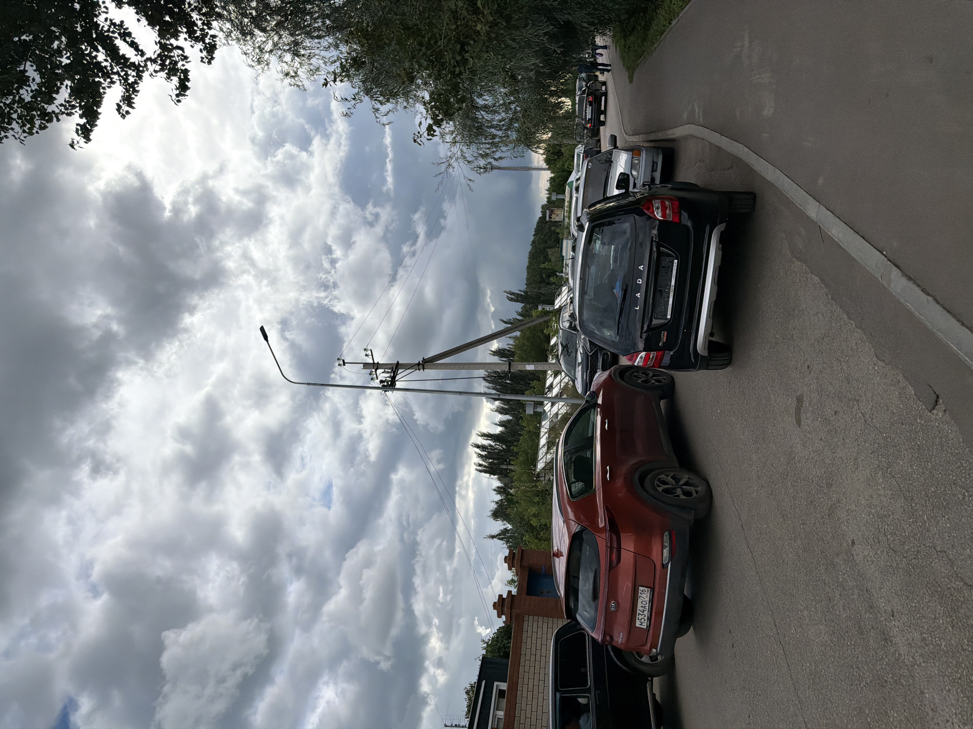 A cloudy day in Yelabuga, Russia, shows a street lined with parked cars. In the foreground, a red Kia Sportage with a license plate M534AO 716 is parked on the left. To its left, a black SUV is partially visible. Behind the Kia, a black Lada Granta is parked. Further down the street, several other cars are parked, including a silver sedan. A sidewalk runs along the right side of the street, with trees and foliage bordering it. A utility pole with wires is visible in the middle ground. The sky is filled with dramatic, dark gray clouds, suggesting impending rain.