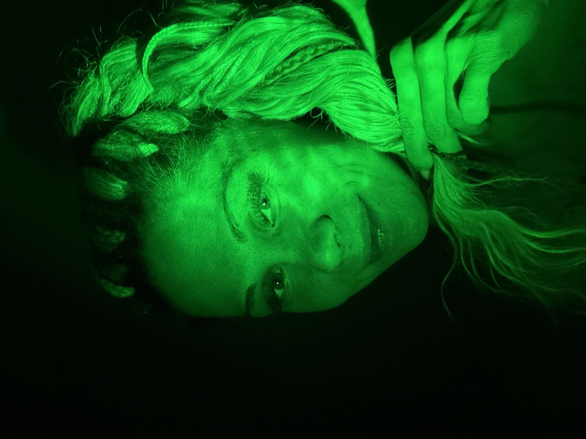 A green-lit portrait of a person is oriented horizontally. The subject has fair skin, light-colored hair styled with braids, and their facial expression appears neutral. A hand is grasping a section of the subject's hair at the top right of the image. The background is black. The location is not discernible.