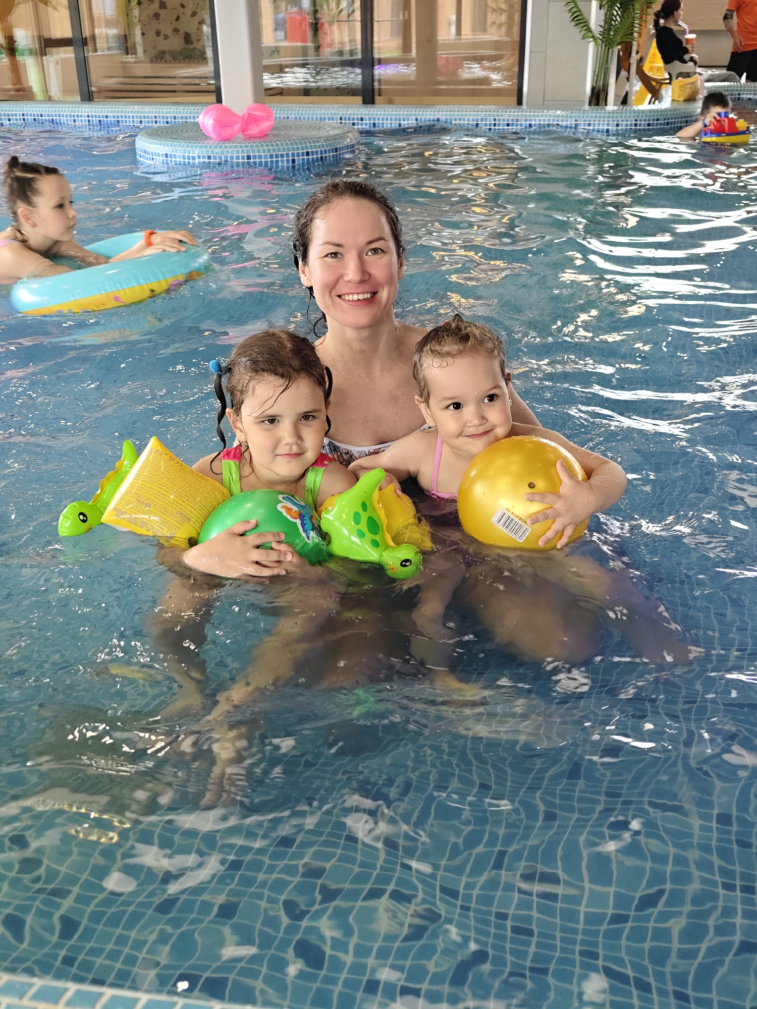 In a brightly lit indoor swimming pool, a woman is standing waist-deep in the water, holding two young children. The pool is lined with small blue and white tiles. The woman has dark hair and is smiling at the camera. One child, on the left, has brown hair in pigtails and is wearing arm floaties. She is holding a green ball. The other child, on the right, has light brown hair and is holding a yellow ball. Another child is in the background with a pool tube, and other people are visible further back. The location is likely a water park in Votkinsk, Russia.