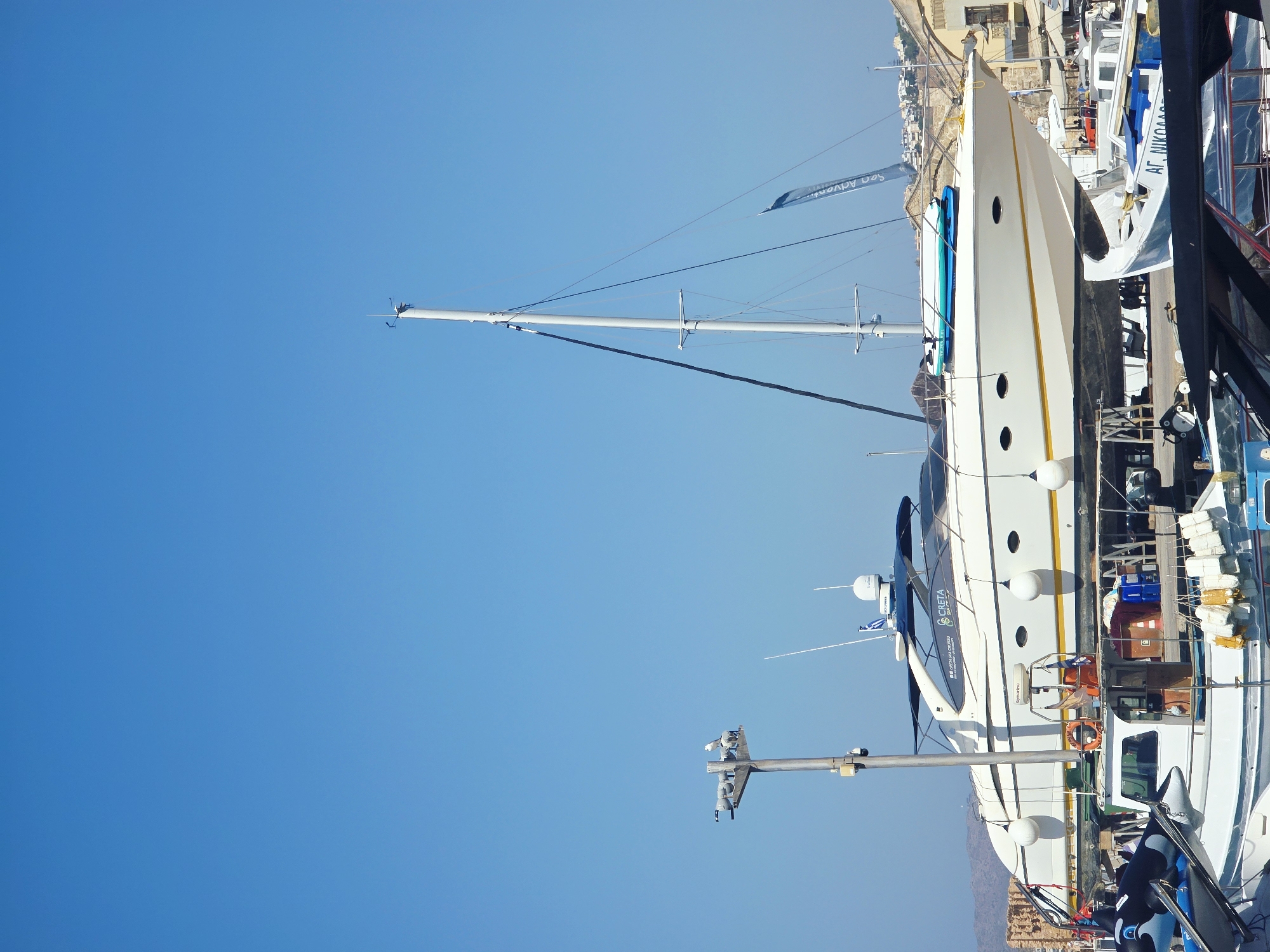 In the city of Chania, Greece, a large white yacht named "CRETA" is dry-docked on a cradle at the harbor. The yacht features a dark blue canopy and a yellow waterline stripe. Below the yacht, another smaller boat is visible. A tall mast with rigging rises behind the yacht, and a blue flag with white lettering, possibly advertising "CRETA SEA CRUISES," hangs from it. In the foreground, a black inflatable orca toy is visible on a boat, and other smaller boats and marine equipment are present in the harbor. The sky is clear and blue, and buildings and hills can be seen in the background.