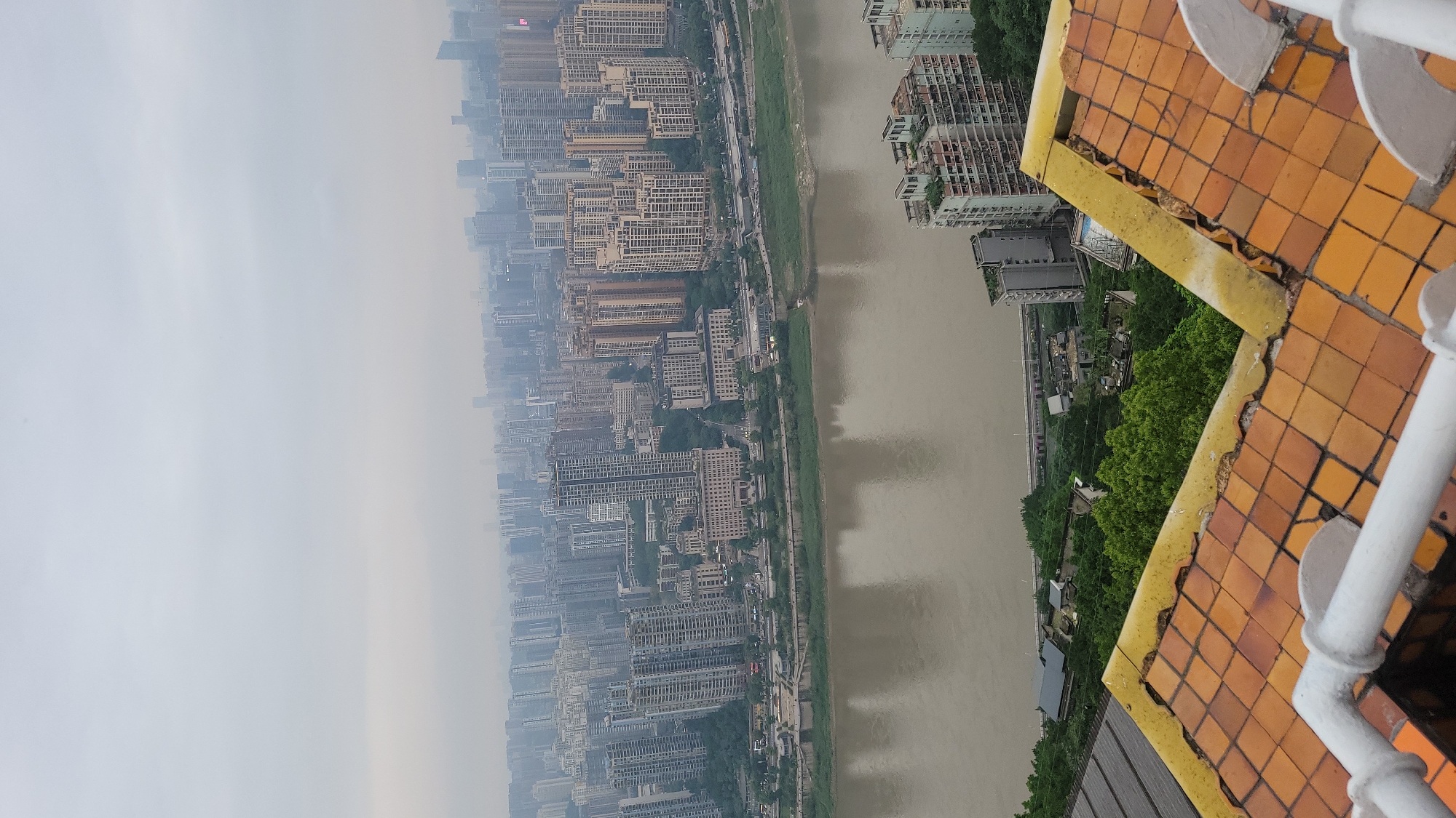 The image shows an aerial view of a cityscape alongside a wide river. The cityscape is densely populated with high-rise buildings, receding into a hazy distance. A river runs parallel to the city, with a green strip of vegetation along its banks. The foreground features part of a rooftop with orange tiles and a white railing.