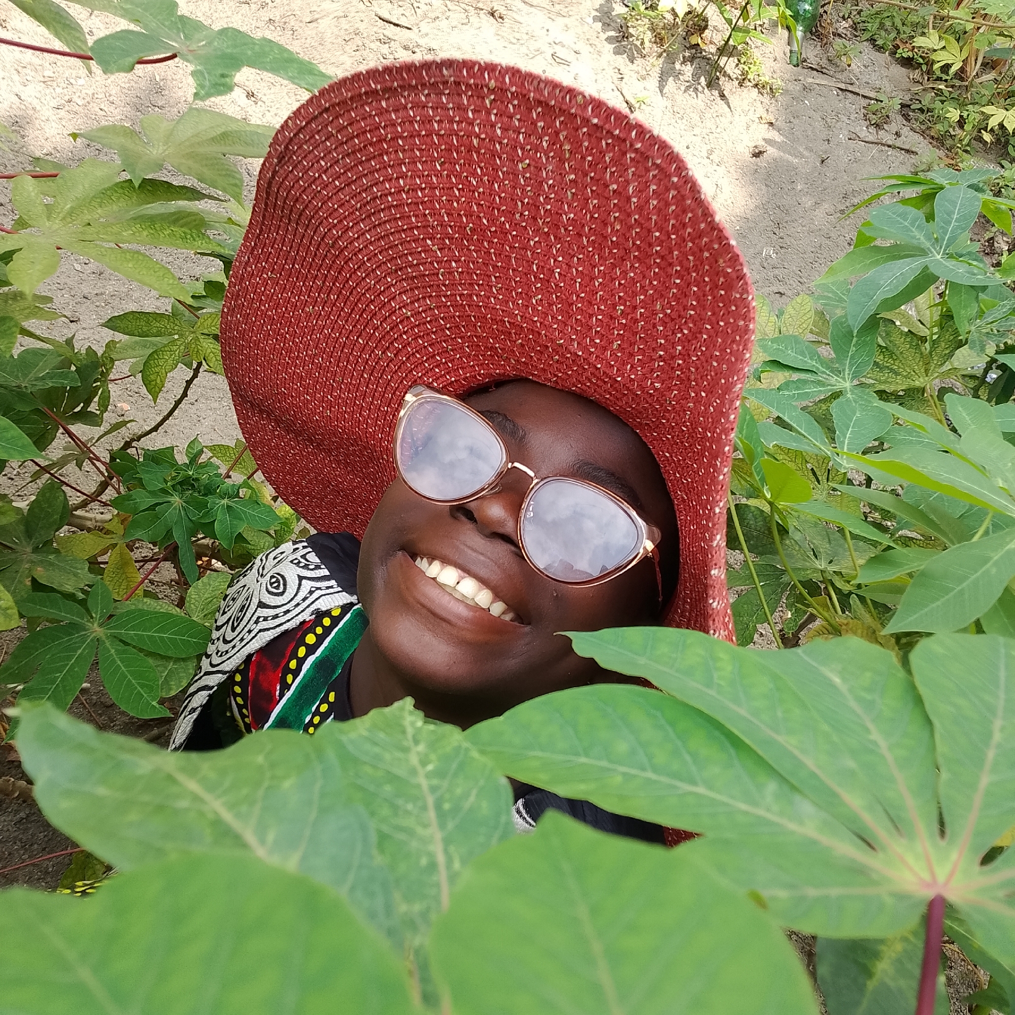 A person wearing a red sun hat and sunglasses smiles broadly, their teeth visible. They are looking upwards and are partially obscured by large green leaves in the foreground. The person is wearing a black garment with a patterned fabric around the neckline. The background appears to be a sandy area with some vegetation. The scene is outdoors, likely in Ado, Nigeria.