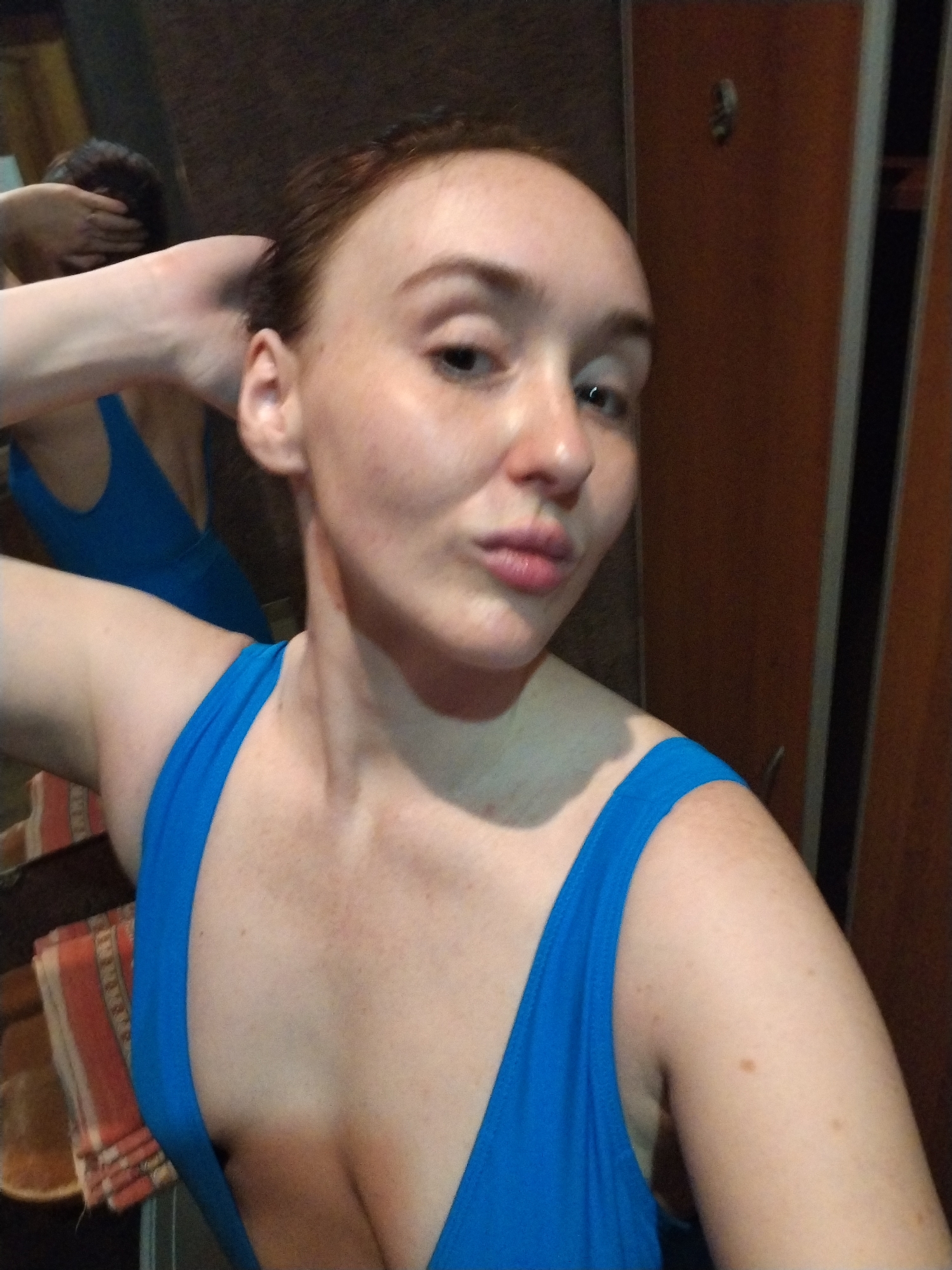 A woman poses for a selfie indoors. She is wearing a blue bathing suit and has red hair. Her right arm is raised with her hand touching her head. A mirror reflects her back. A towel is visible on the left, and a wooden cabinet with metal accents is on the right. The location is Chaikovsky, Russia.