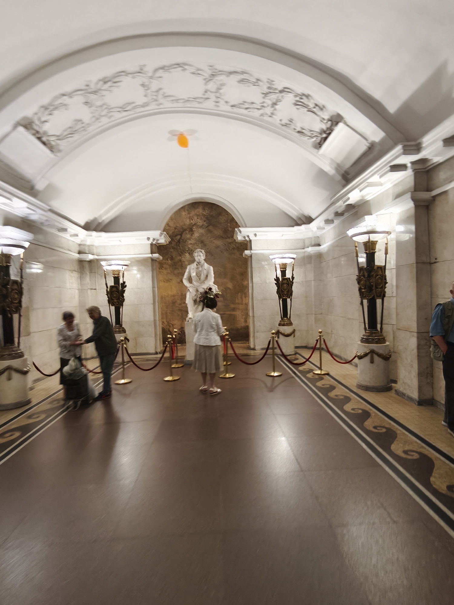 This is a photograph of the interior of the metro station "Pushkinskaya" in Saint Petersburg, Russia. The station's architecture is characterized by high vaulted ceilings with decorative stucco work and marble walls. A large white statue of Alexander Pushkin stands in an alcove at the far end of the platform. Two floor lamps, adorned with ornate metalwork, flank the statue. Red velvet ropes on gold stanchions demarcate areas of the platform. Several people are visible: two individuals appear to be talking near the entrance, one person is standing before the statue, and a person in a blue shirt and vest is partially visible on the right side of the frame. The floor is polished and reflects the light from the lamps and the ceiling. An orange balloon is suspended from the ceiling.
