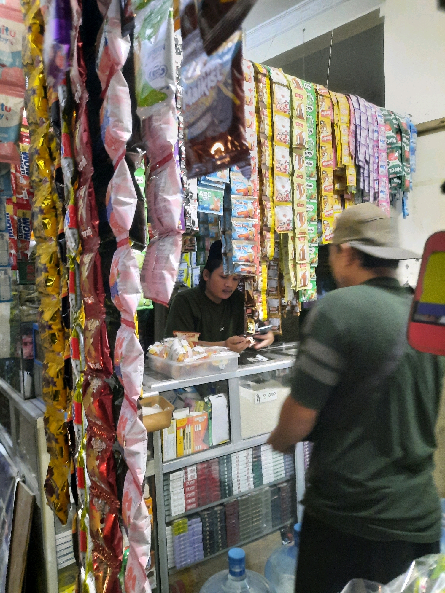 The image depicts the interior of a small convenience store in East Jakarta, Indonesia. The store is densely stocked with various items, primarily packaged snacks and beverages hanging from the ceiling and displayed on shelves. In the foreground, a person wearing a dark green shirt and a light brown hat is partially visible from the back, facing away from the camera. This individual appears to be interacting with the store owner, who is a young woman sitting behind a counter, wearing a dark green t-shirt and a black headscarf. She is looking down, possibly at a transaction or a smartphone.
The shelves behind the counter are filled with rows of packaged goods, including what appear to be cigarettes and other small items. A price of "Rp 13.000" is visible on a piece of paper or sign on the counter. Numerous colorful, foil-wrapped snack packages hang in rows, creating a vibrant and cluttered visual. The lighting suggests an indoor environment, possibly illuminated by artificial light sources. The overall impression is that of a typical neighborhood shop in Indonesia.