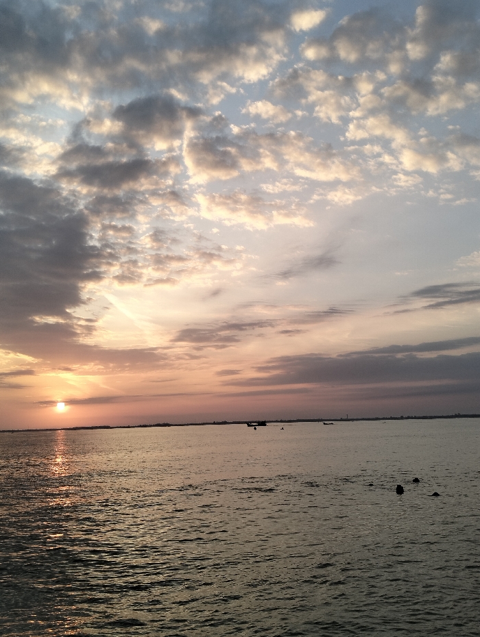 A wide expanse of water reflects the light of a setting sun. The sky is a blend of soft blues, grays, and pinks, filled with scattered clouds. A few small boats are visible on the horizon. The water's surface has gentle ripples. Dark objects, potentially swimmers or debris, are scattered in the foreground. The overall scene suggests a serene and peaceful moment at dusk.