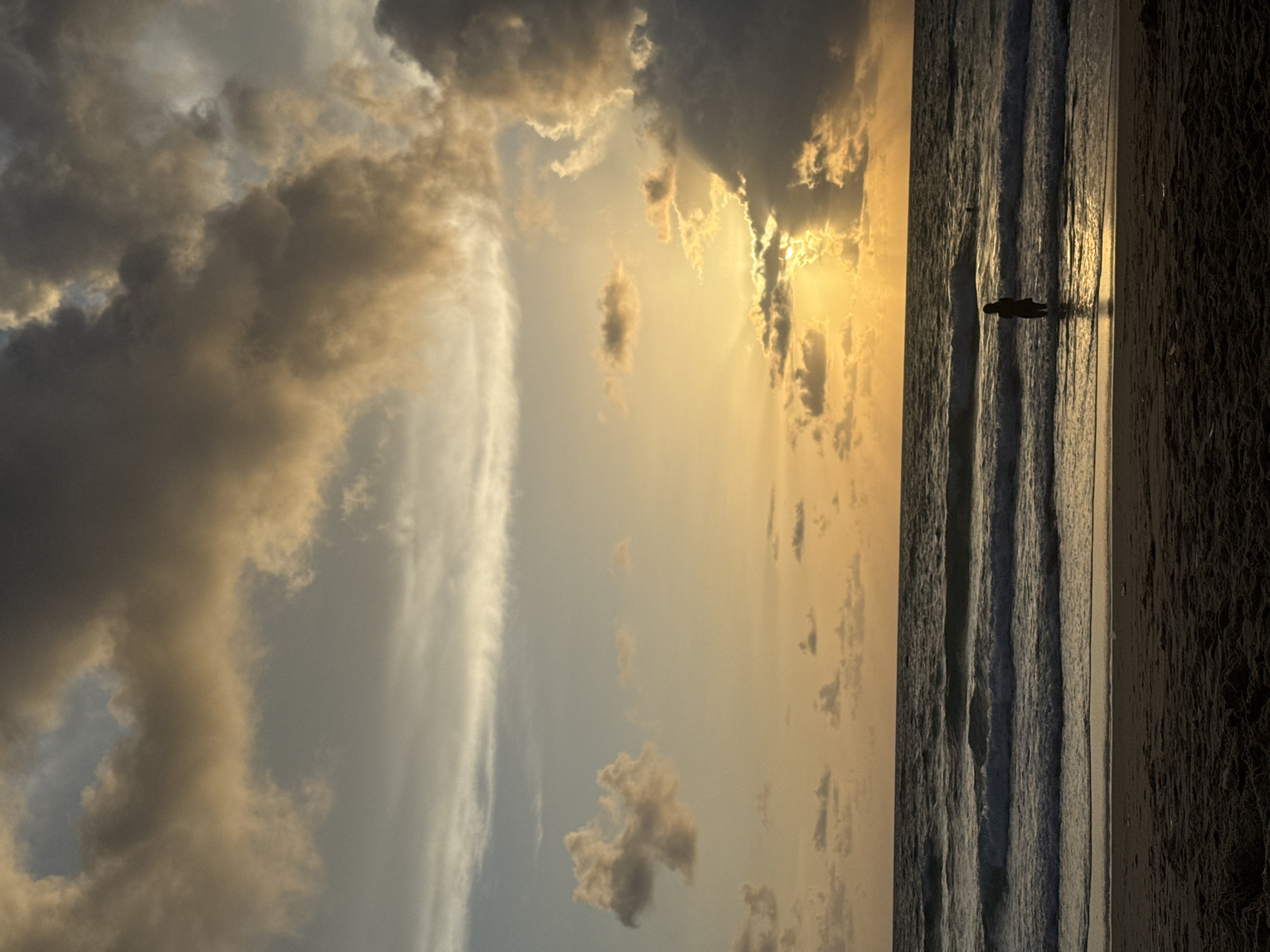 A person stands in the shallow water at Karon Beach, Thailand, silhouetted against a dramatic sunset. The sky is filled with heavy, dramatic clouds tinged with gold and orange as the sun begins to set, with rays of light piercing through gaps in the clouds. The ocean is calm with gentle waves rolling towards the shore. The beach in the foreground is dark and sandy, with the texture of wet sand visible where the waves recede. Another person can be seen further out in the water.