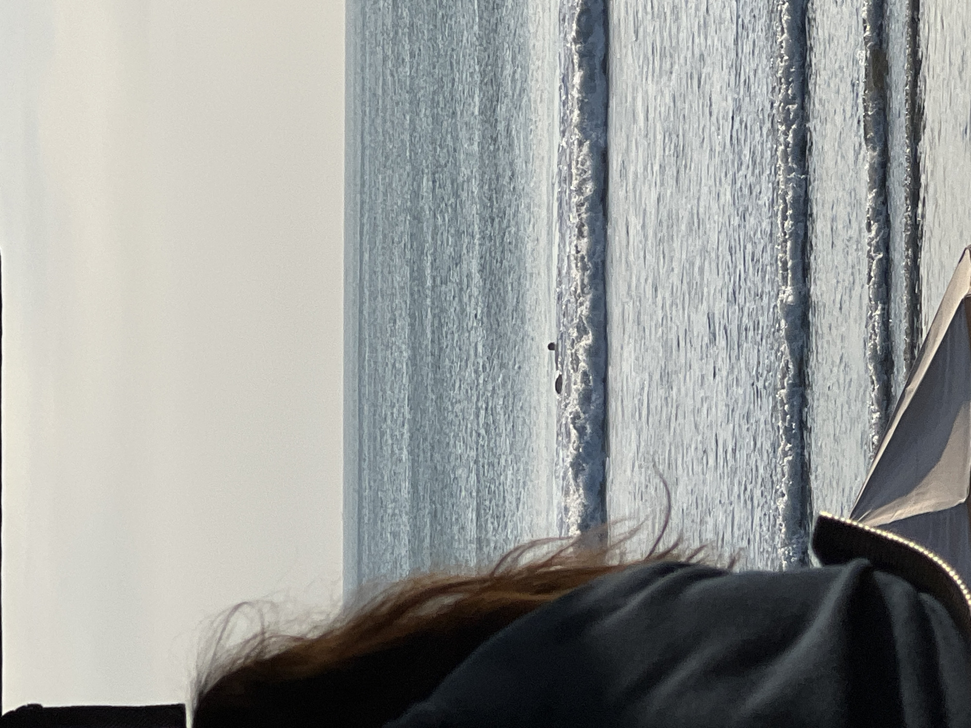 A person, seen from behind with reddish-brown hair and wearing a dark hoodie, sits in the foreground, looking out at the ocean. The ocean is calm with gentle waves breaking on the shore. In the distance, a lone figure can be seen in the water, possibly a surfer or swimmer. The sky is overcast, with a hazy sun. The scene appears to be a beach in Kuta, Indonesia.