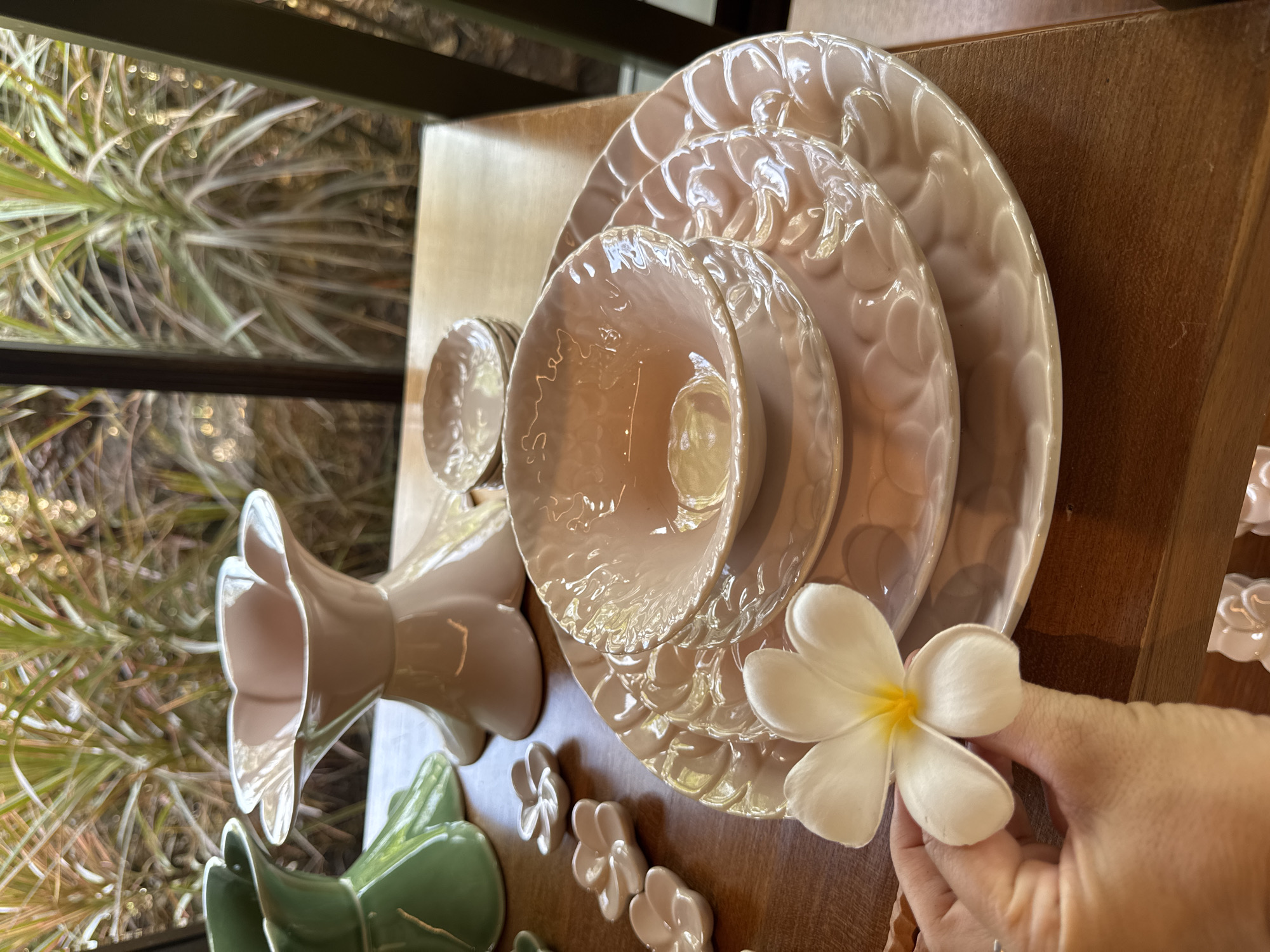 A still life is arranged on a wooden surface. The composition includes stacked, light pink ceramic plates and bowls, a vase shaped like a flower, and small ceramic flower-shaped ornaments. A white plumeria flower with a yellow center is held in a person's hand. The scene suggests a retail or display setting, possibly related to home decor or tableware. The image originates from Kuta Selatan, Indonesia.