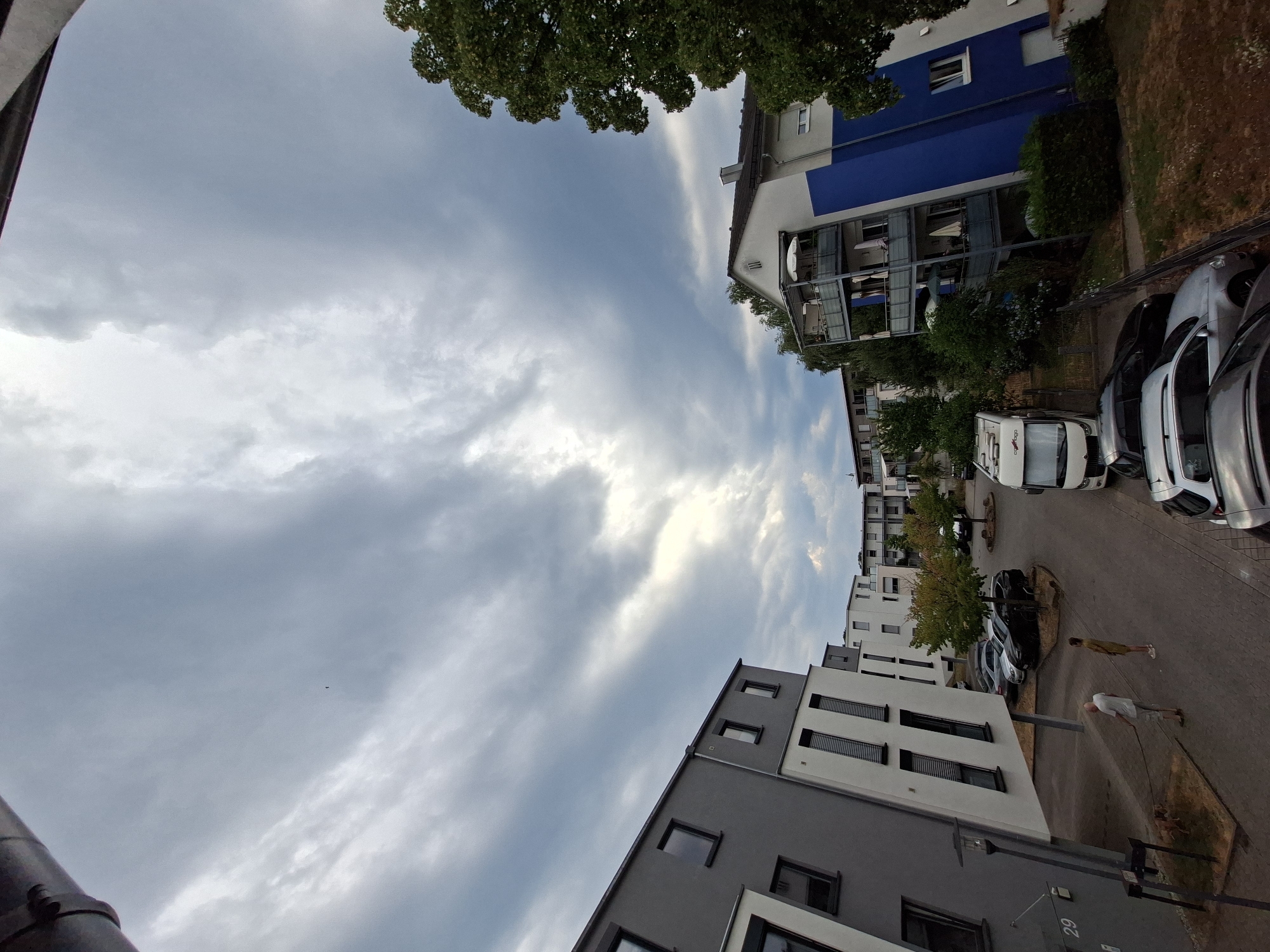 The image shows an outdoor scene in Baden-Baden, Germany, likely a residential complex. The perspective is from an elevated viewpoint, looking down into a courtyard area. In the foreground, on the left, is a modern apartment building with grey and white facades and numerous windows. To the right of this building, a small dog on a leash is being walked by a person wearing a white t-shirt and light-colored shorts. Another person in a yellow dress stands nearby. The courtyard is paved with interlocking stones. Several cars, including a white camper van and dark-colored sedans, are parked in the area. Further into the scene, another residential building with blue and white sections and balconies is visible, along with trees and other apartment buildings in the background. The sky is heavily clouded, with patches of bright light suggesting the sun is obscured. The overall atmosphere appears overcast.