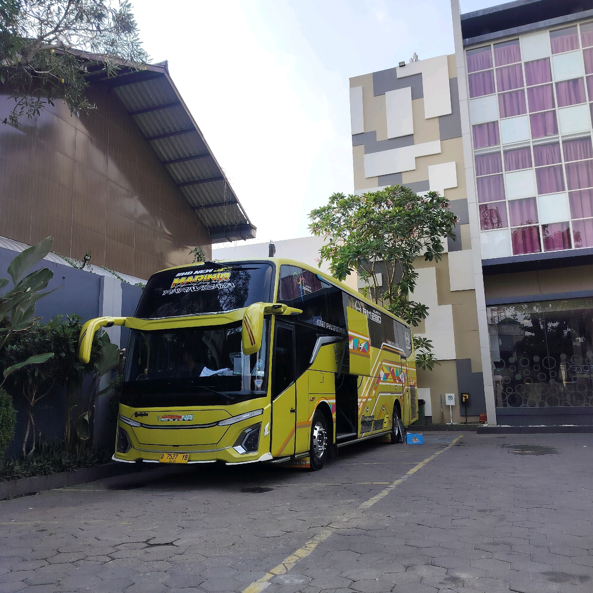 In Yogyakarta, Indonesia, a bright yellow double-decker bus is parked in front of a modern building with a patterned facade and large windows with purple curtains. The bus, labeled "SHO NEW MARNIR" and "BUS PARIWISATA," has intricate colorful designs along its side. In front of the bus, a license plate reads "D 7537 YB." The ground is paved with hexagonal stones, and there are some patches of wetness. To the left of the bus, a wall is covered in dark siding, with lush green plants growing around it. A tree with small pink flowers is visible between the bus and the building. The sky is bright and overcast.