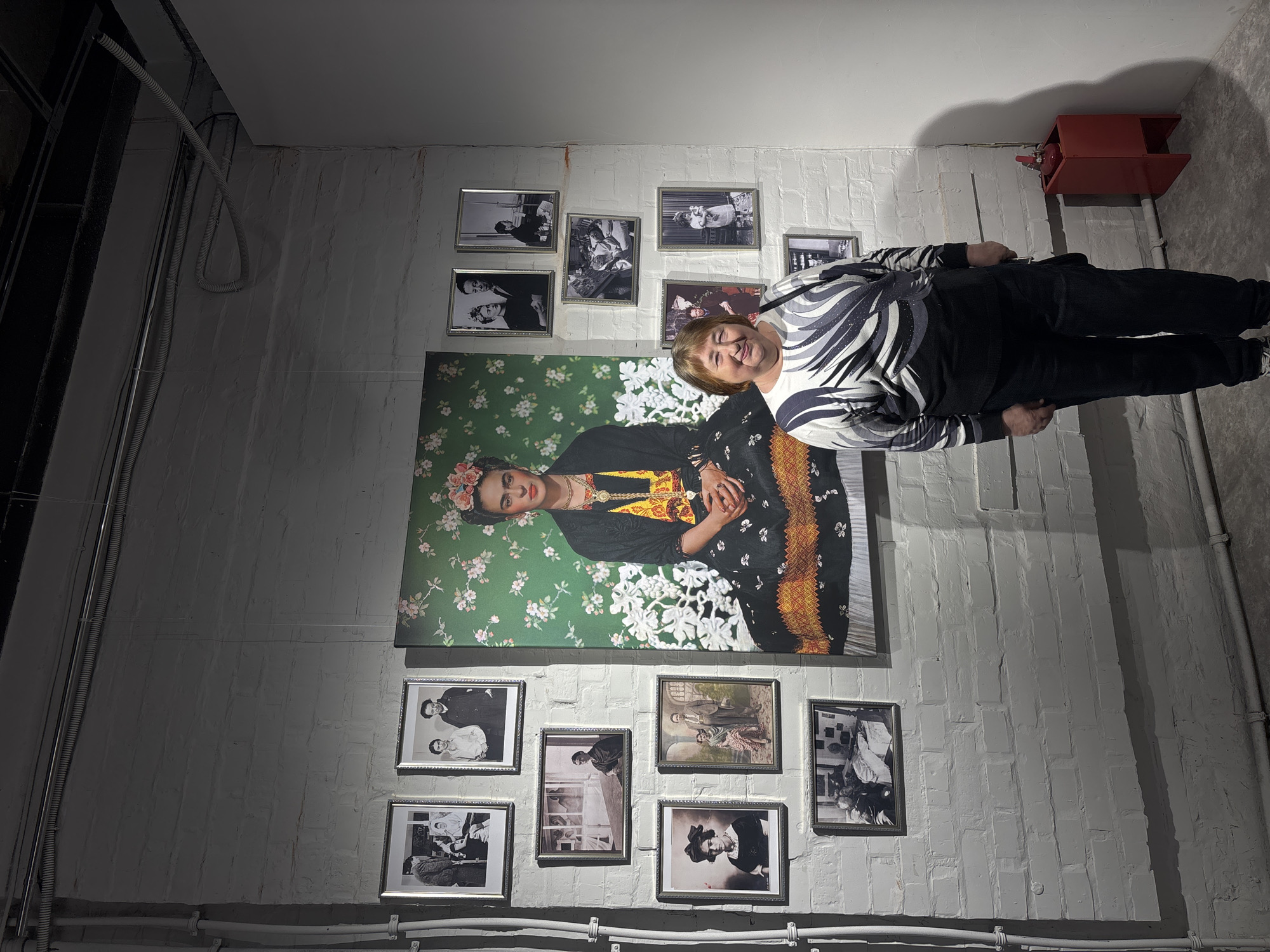 The scene is an indoor setting, possibly a basement or gallery space, in Krasnoyarsk, Russia. The walls are made of white brick. A woman in a black and white patterned sweater and dark pants is standing facing the camera. Behind her is a large framed portrait of Frida Kahlo with a green floral background. Surrounding the portrait are several smaller framed black and white photographs. A red fire extinguisher is mounted on the wall in the upper right corner. There are also visible conduits and wiring along the walls and ceiling. The lighting is uneven, with some areas brighter than others. The scene appears to be a display or exhibition of photographs and art.
