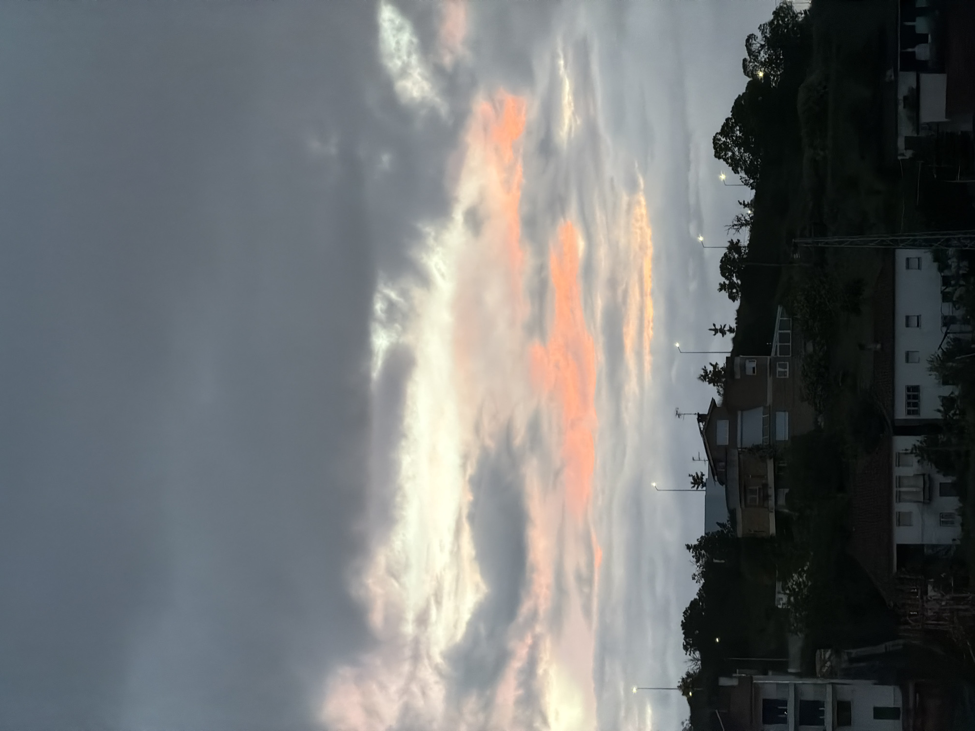 The image shows a view in Bragança, Portugal at dusk. The sky is predominantly overcast with gray clouds, but some areas are illuminated with orange and white from the setting sun. On the right side of the frame, residential buildings are visible along a hillside, interspersed with trees and streetlights. Some buildings are white with visible windows, and others have darker facades.