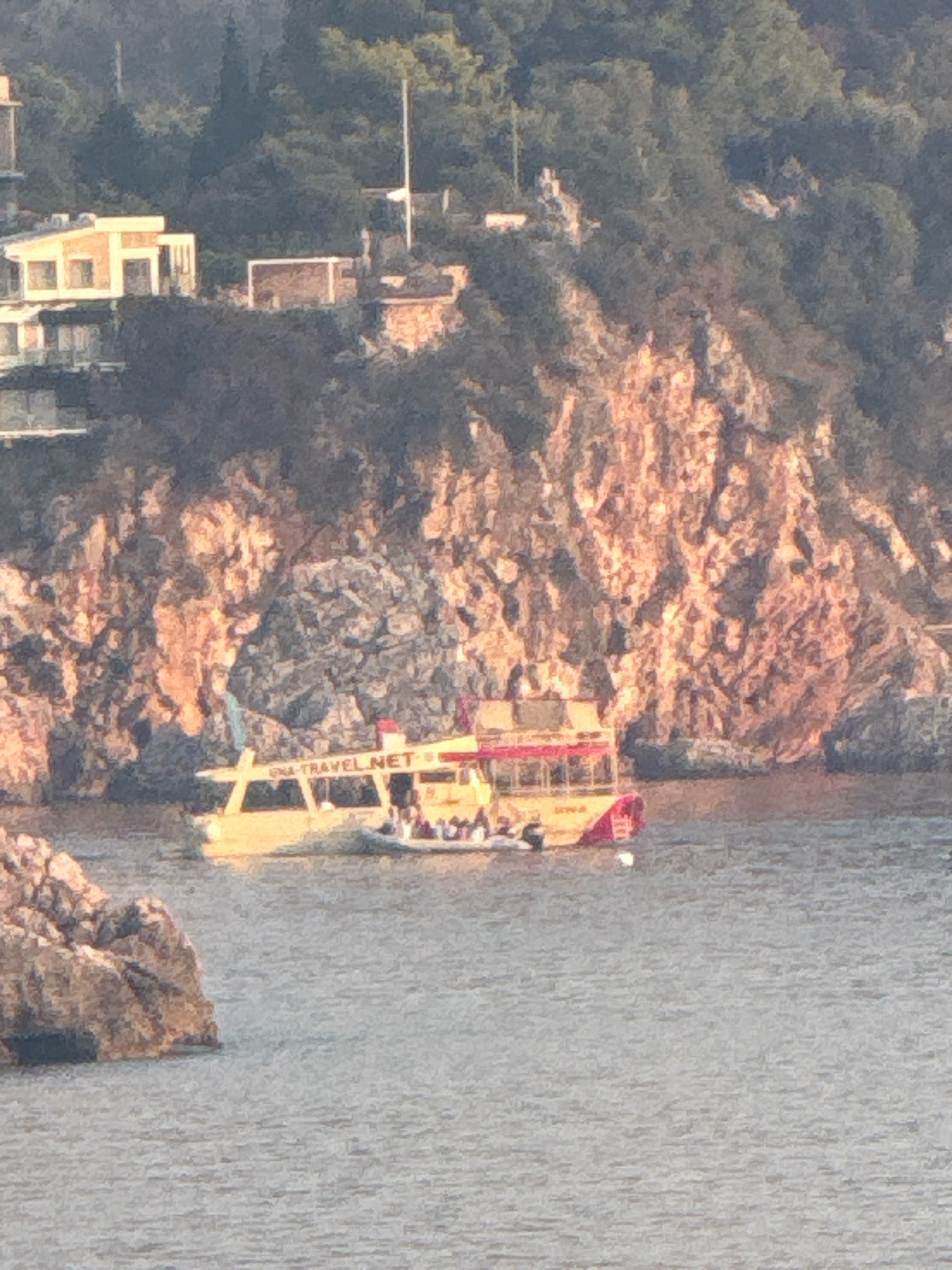 The image shows a tour boat with people on board sailing near a rocky coastline with buildings on top of the cliffs. The boat is yellow with a red top and has "EPMARAVEL.NET" written on its side. The coastline is covered with trees and rocks, and the buildings appear to be hotels or residences. The sea is calm and reflects the sunlight. The location is Kamenovo, Montenegro.