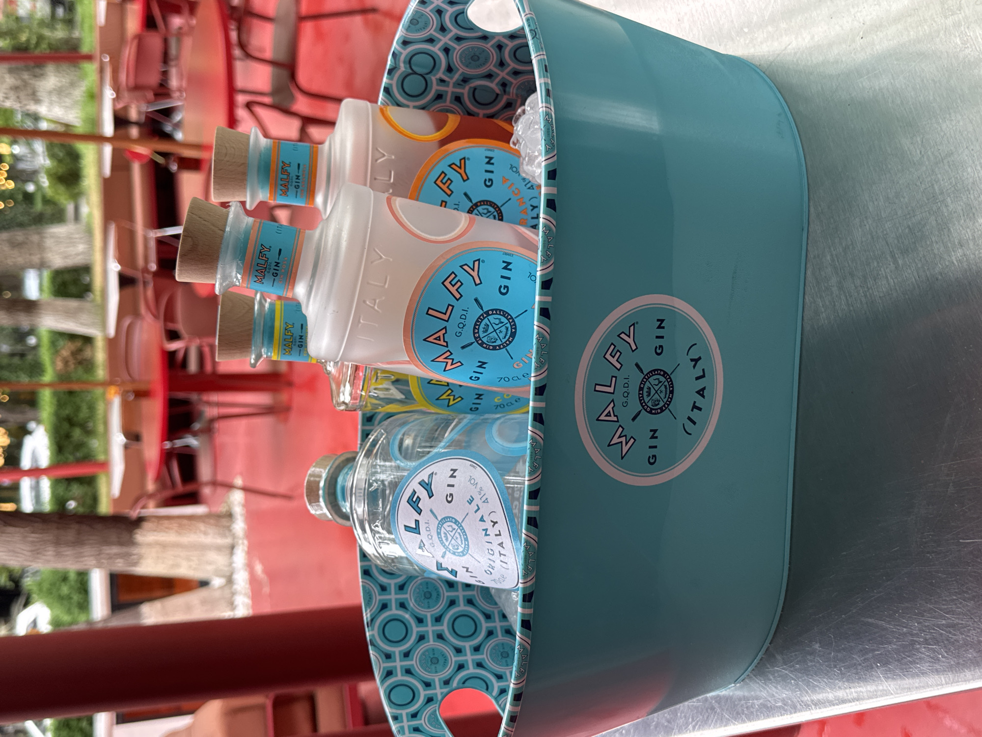 A portrait-style, outdoor shot depicts a light blue, oval-shaped bucket filled with ice and at least four bottles of "Malfy Gin" from Italy. The bucket has two circular holes near the top of each end, apparently used for carrying. A repeating light blue and dark blue circular design lines the inside of the bucket. "MALFY" in a large blue font with an emblem is on the side of the bucket. Each bottle is clear glass with a round wooden cork and a blue label on the front. The bottles are filled with varying shades of clear and light yellow liquid. The background contains red metal chairs and a table, along with blurred greenery and trees, suggesting an outdoor patio or restaurant setting. The bottles are close-up, suggesting they are the subject of the image. There is no evidence of the city of Yerevan, Armenia, or any other people.