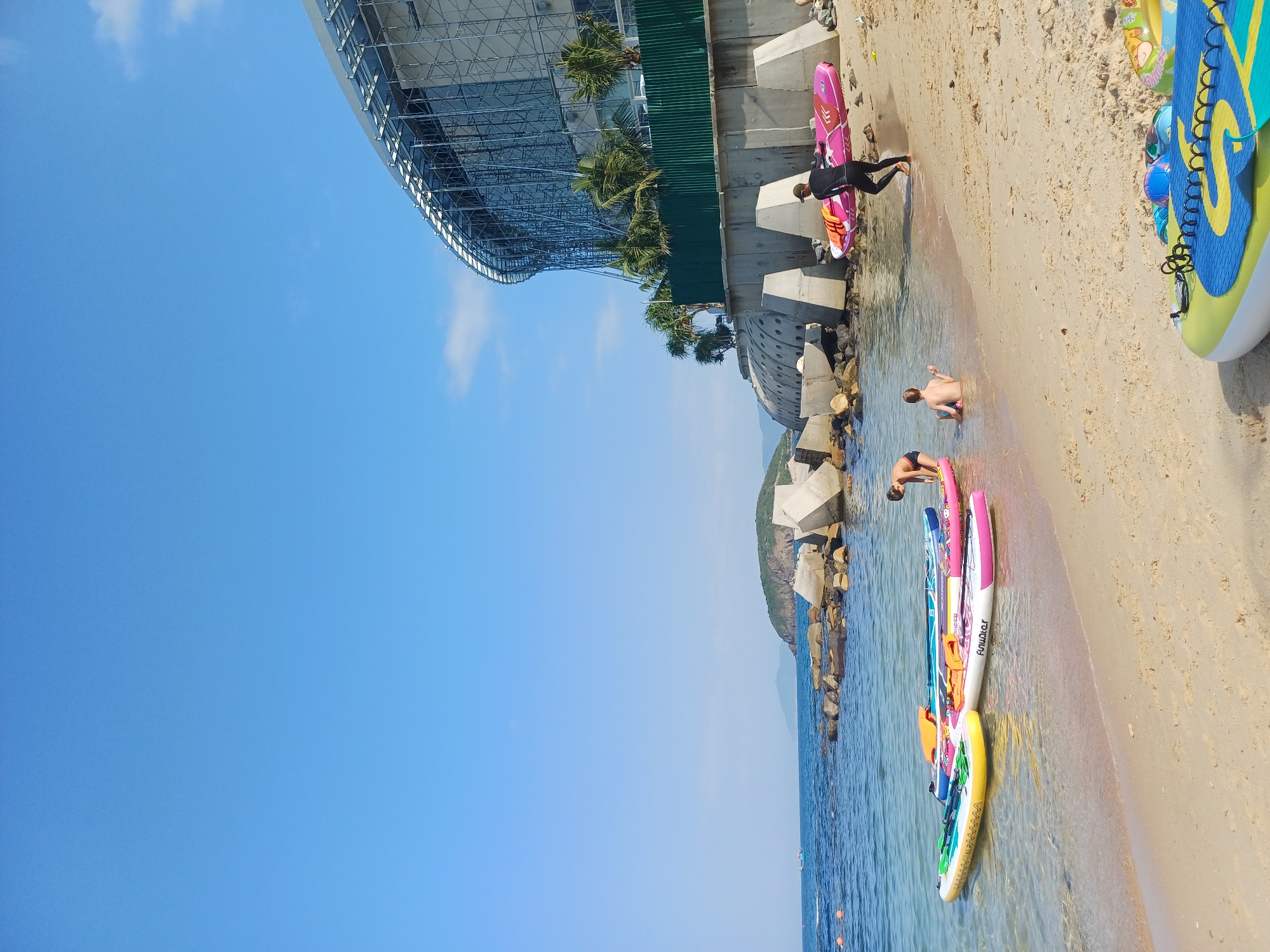 In Nha Trang, Vietnam, a sunny beach scene unfolds by the ocean. Several inflatable stand-up paddleboards, in vibrant yellow, pink, and blue, are partially submerged in the clear, shallow water near the shore. On the sandy beach, a person wearing a black wetsuit carries a pink paddleboard, walking away from the water. Two children are visible near the water's edge; one is sitting in the shallow water, and the other is standing, looking towards the paddleboards.
In the background, large, precast concrete blocks form a coastal barrier, with a modern building under construction featuring a complex scaffolding structure behind a green fence. Palm trees are interspersed along the embankment. The calm ocean stretches to the horizon, meeting a clear blue sky with a few wispy clouds. The overall impression is a recreational beach area adjacent to a construction site.