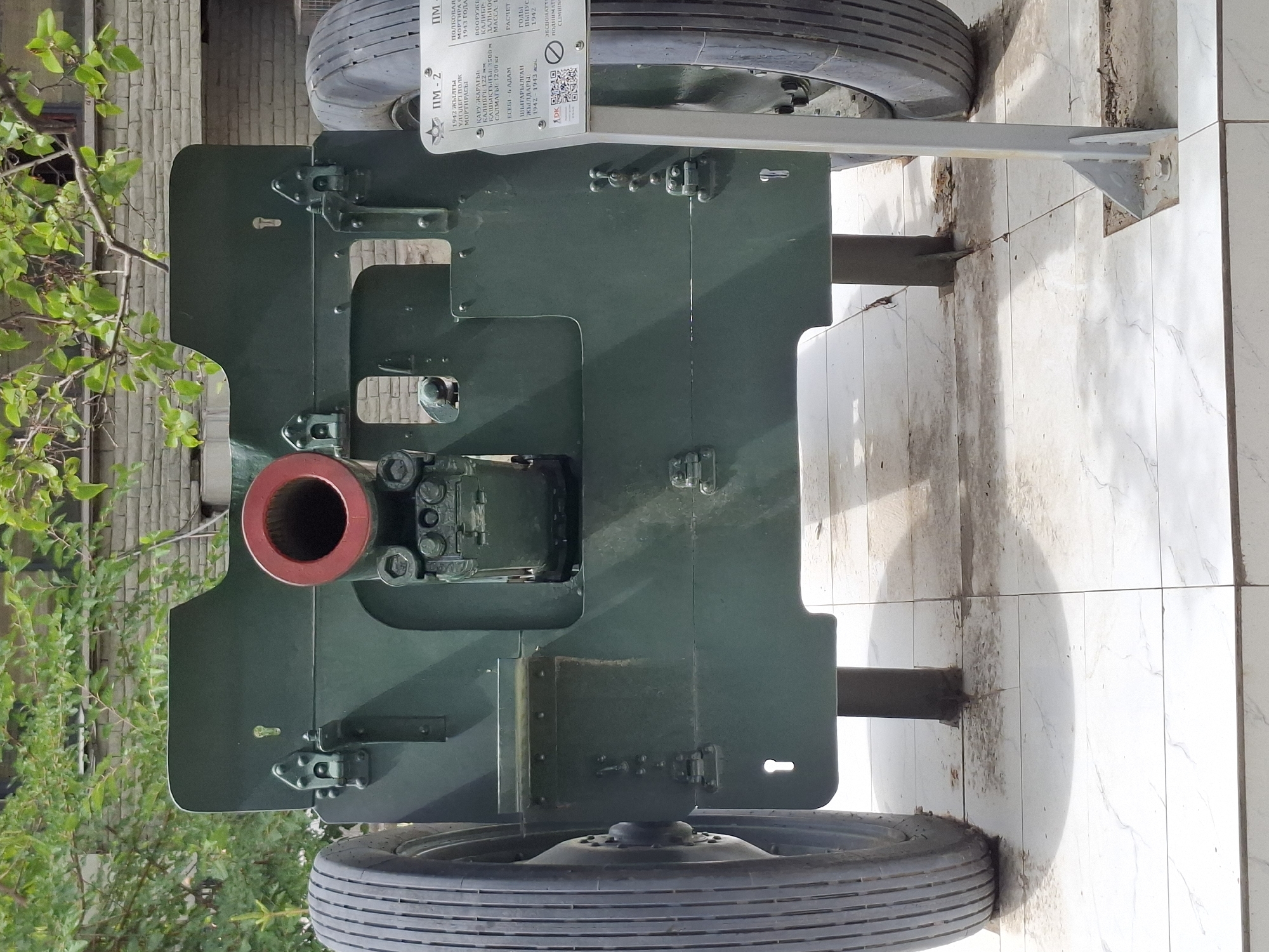 A 76mm divisional gun M1939 (3-76) is on display outdoors, positioned on a tiled ground. The gun is painted dark green and features large pneumatic tires. A placard with information about the artillery piece is visible to the right of the gun, displaying Cyrillic text and a QR code. The background consists of a brick wall and green foliage from trees. The overall setting appears to be a museum or memorial site in Almaty, Kazakhstan, given the style of the gun and the presence of a multilingual placard, which is common in such locations.