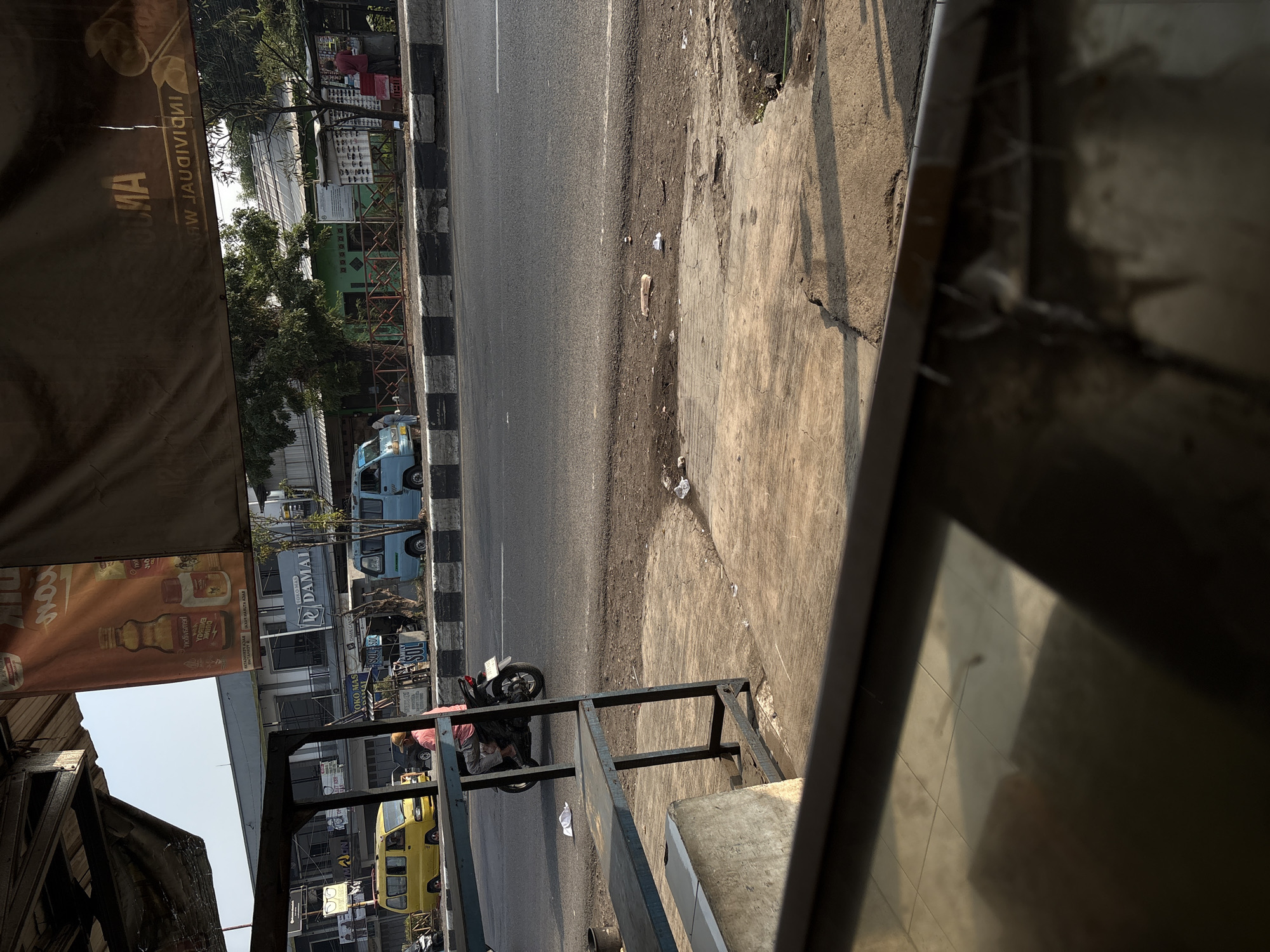 The image depicts a street scene in Palimanan, Indonesia. In the foreground, a structure with blue metal shelving is partially visible. Beyond this, a paved road runs horizontally across the frame, with a black and white striped median. A motorcycle with a rider wearing a pink shirt and grey pants is traveling on the road. To the left, a yellow minivan is parked. Further down the road, a light blue van is also visible. Buildings line the right side of the road, with one appearing to be a shop with a sign that reads "Toko Mas Damai" (Jewelry Store Damai). There are trees and a fence in front of the buildings. A banner with Indonesian text is hanging above the street. The environment appears to be a commercial area with moderate traffic. The lighting suggests it is daytime, possibly late afternoon due to the shadows.