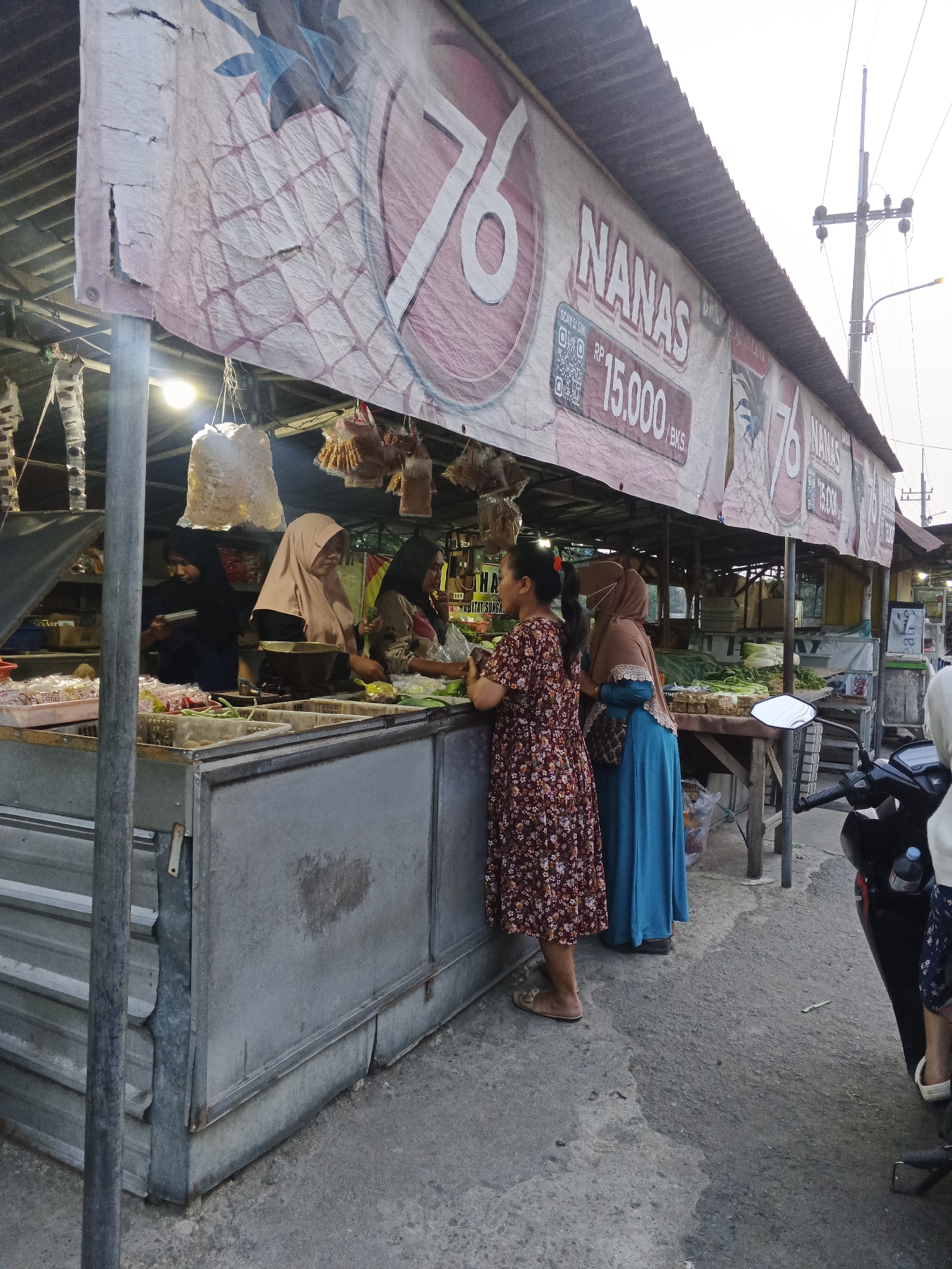 The image depicts an outdoor market scene in Penambangan, Indonesia. In the foreground, a market stall displays produce, including green vegetables. Several individuals are interacting at the stall; two women in hijabs are attending to customers, while two other women, one in a floral dress and the other in a blue hijab and dress, appear to be making purchases. A motorcycle is parked to the right of the frame. A banner overhead advertises "NANAS" (Pineapple) for "15.000 / BKS." The market appears to be set up along a street, with a power pole and lines visible in the background. The overall atmosphere suggests a typical daytime market environment.