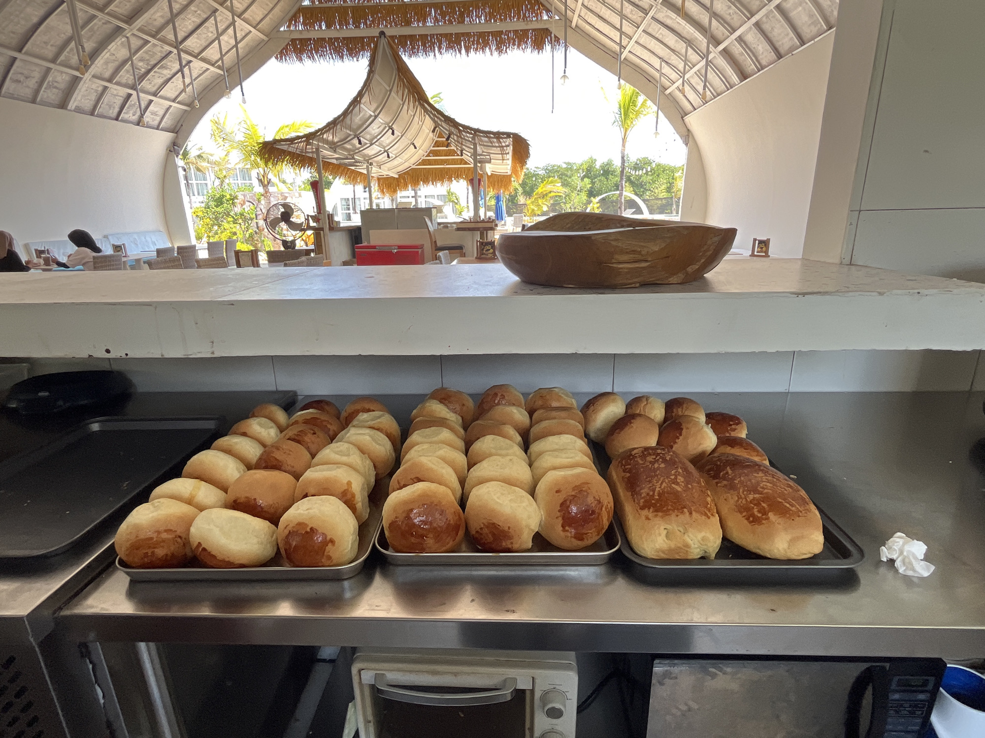 A high-angle, medium shot captures a buffet-style food preparation area, likely in Kemloko, Indonesia, given the context. In the foreground, a stainless steel countertop is laden with two metal trays filled with freshly baked bread rolls. The rolls are varied in color, ranging from pale golden to rich brown, indicating different baking times or ingredients. To the right of the rolls, a single loaf of bread, also golden brown, rests on a dark metal tray. To the left of the trays, a black rectangular object is partially visible, possibly a serving dish or equipment. Beneath the countertop, a small white toaster oven is situated.
The background reveals an open-air dining area with a distinctive thatched roof structure supported by white beams, suggesting a tropical or beachside location. Sunlight filters through the roof, illuminating the space. Palm trees and lush greenery are visible beyond the covered area, hinting at an outdoor resort or restaurant setting. Two individuals are faintly visible in the distance, seated at a table. The overall impression is one of a serene and well-prepared dining establishment.