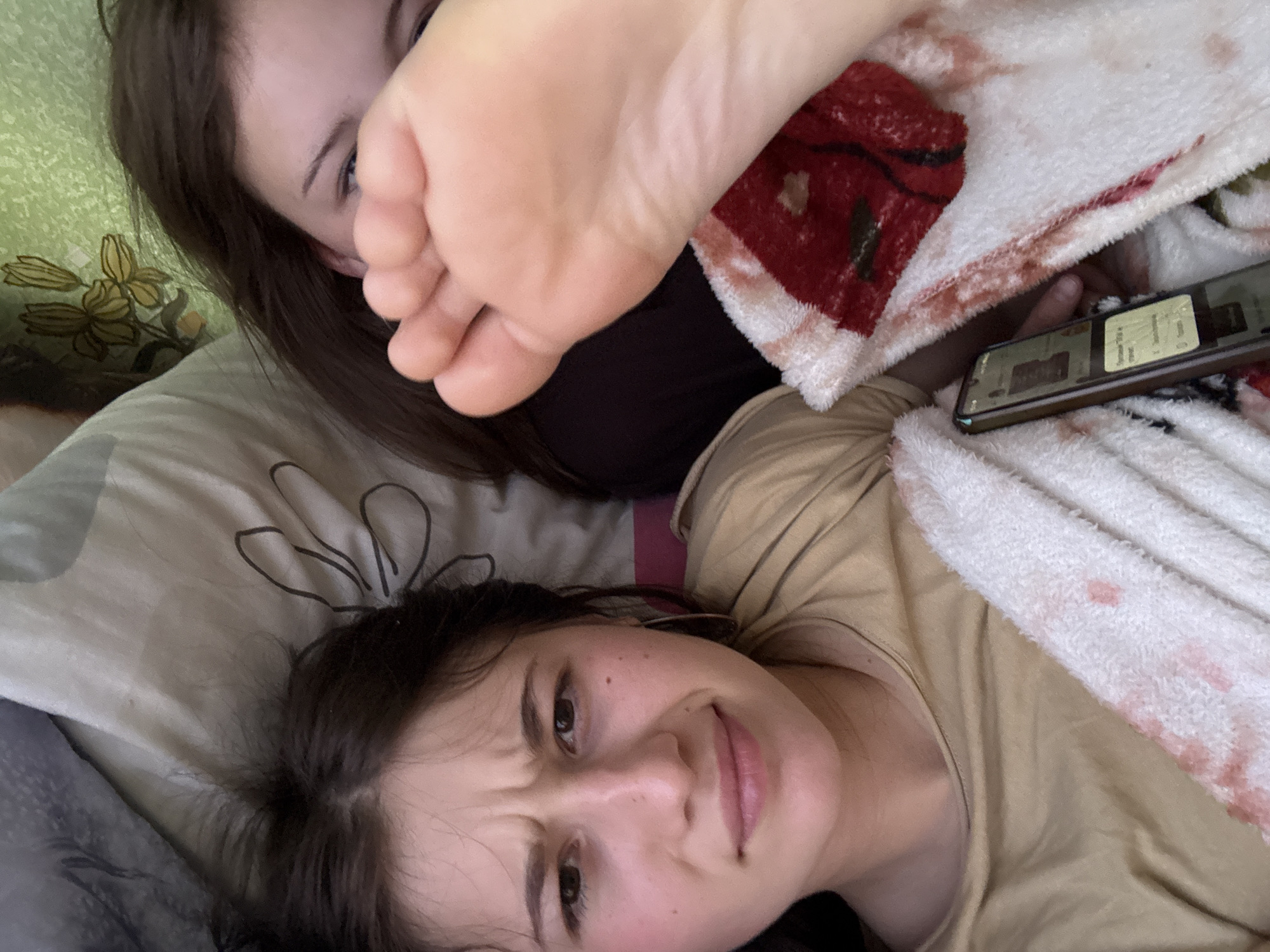 This intimate, top-down shot captures two young women in what appears to be a bedroom setting in Фоки, Russia. The image is taken at an angle, suggesting a casual, personal perspective, possibly a selfie.
In the upper left, one woman's face is partially obscured by a bare foot, likely her own, with only her dark hair and one eye visible. The foot, with its sole facing the camera, takes a prominent central position, adding a playful and slightly intrusive element to the scene. She is lying on a light-colored pillow, next to a green wall or headboard with a yellow floral pattern.
In the lower right, the second woman lies on her side, looking directly at the camera with a scrunched-up facial expression, suggesting playful annoyance or a "what are you doing?" look. Her dark hair is spread around her head on a beige pillow, and she wears a light-colored top.
Between them, a white blanket with red splatters or a distressed pattern is visible. A smartphone rests on this blanket, displaying what looks like a music streaming app, with text possibly indicating "Spotify Free" and UI elements. The overall atmosphere is one of relaxed, informal interaction and lighthearted teasing between the two individuals, likely friends or family, enjoying a moment indoors.