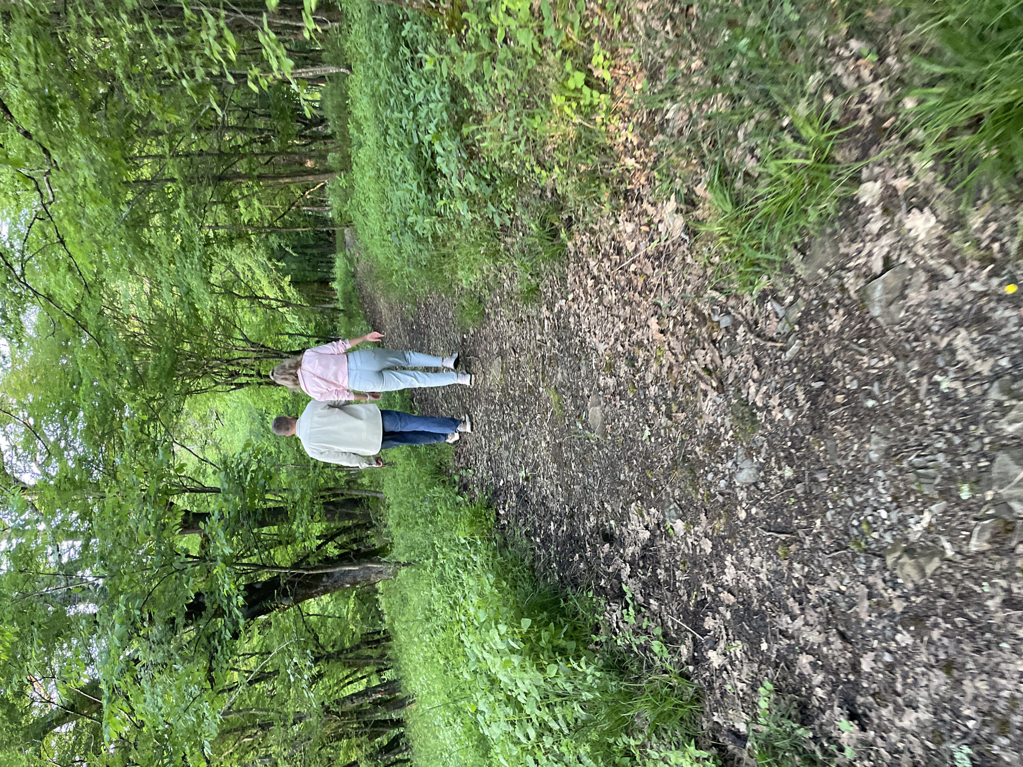 Two people are walking away from the viewer on a narrow path in a wooded area. The path is covered with leaves and dirt. Trees line both sides of the path, with dense green foliage. The person closer to the camera is wearing a light-colored jacket and blue jeans. The other person is wearing a pink shirt and blue jeans. The location appears to be a natural area, possibly a park or forest. There is no evidence to indicate this photo was taken in Olginka, Russia.